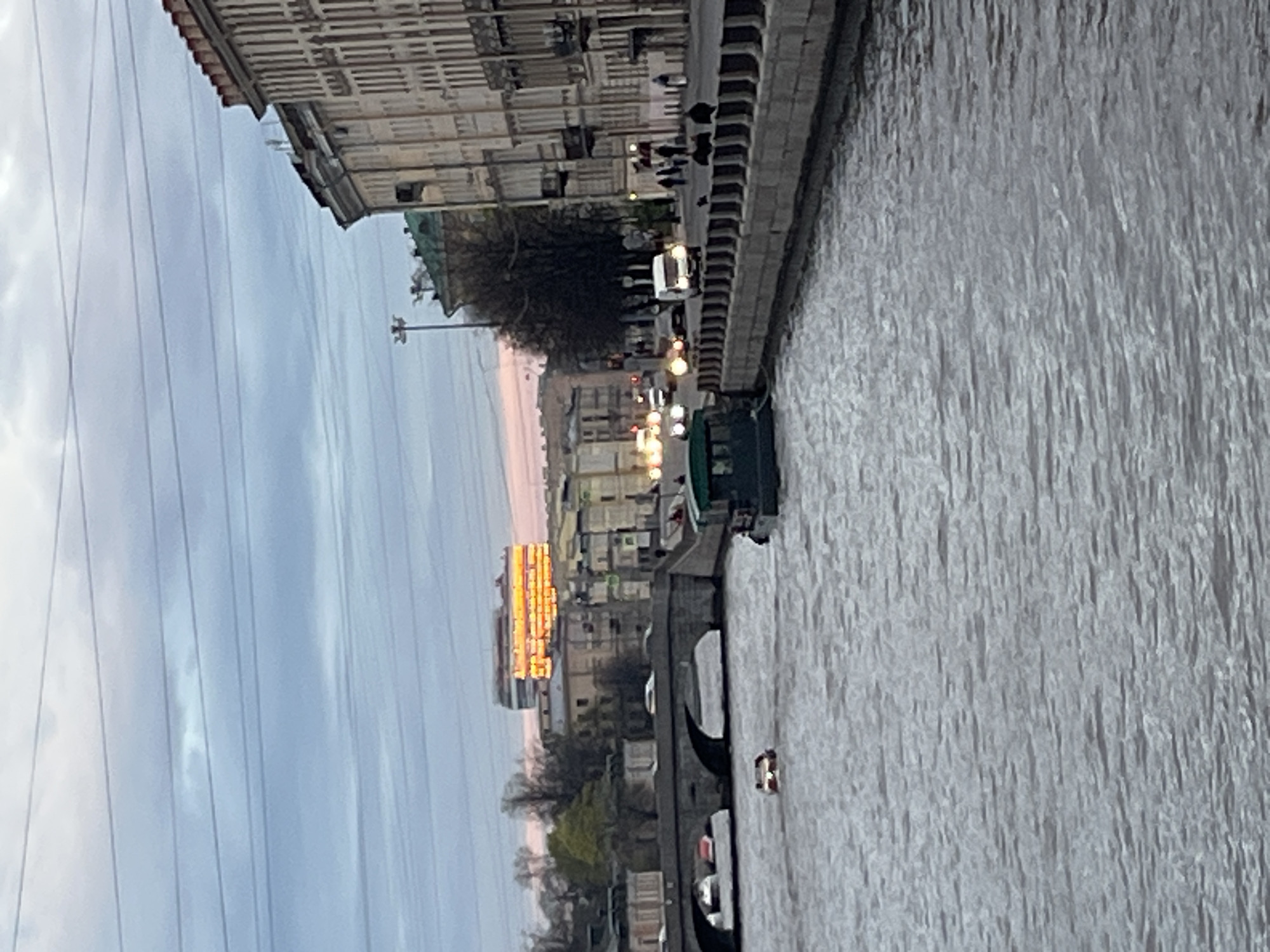 This image depicts a view of the Griboyedov Canal in Saint Petersburg, Russia. The canal is in the foreground, with a dark-colored boat on the water. The canal's embankment is visible on the right side of the frame. Buildings are on the left side of the frame, with some people visible along the embankment. A building with yellow lighting is further in the background, and a bridge with boats passing beneath it is visible in the middle ground. The sky is overcast, with power lines prominent in the upper-left corner.