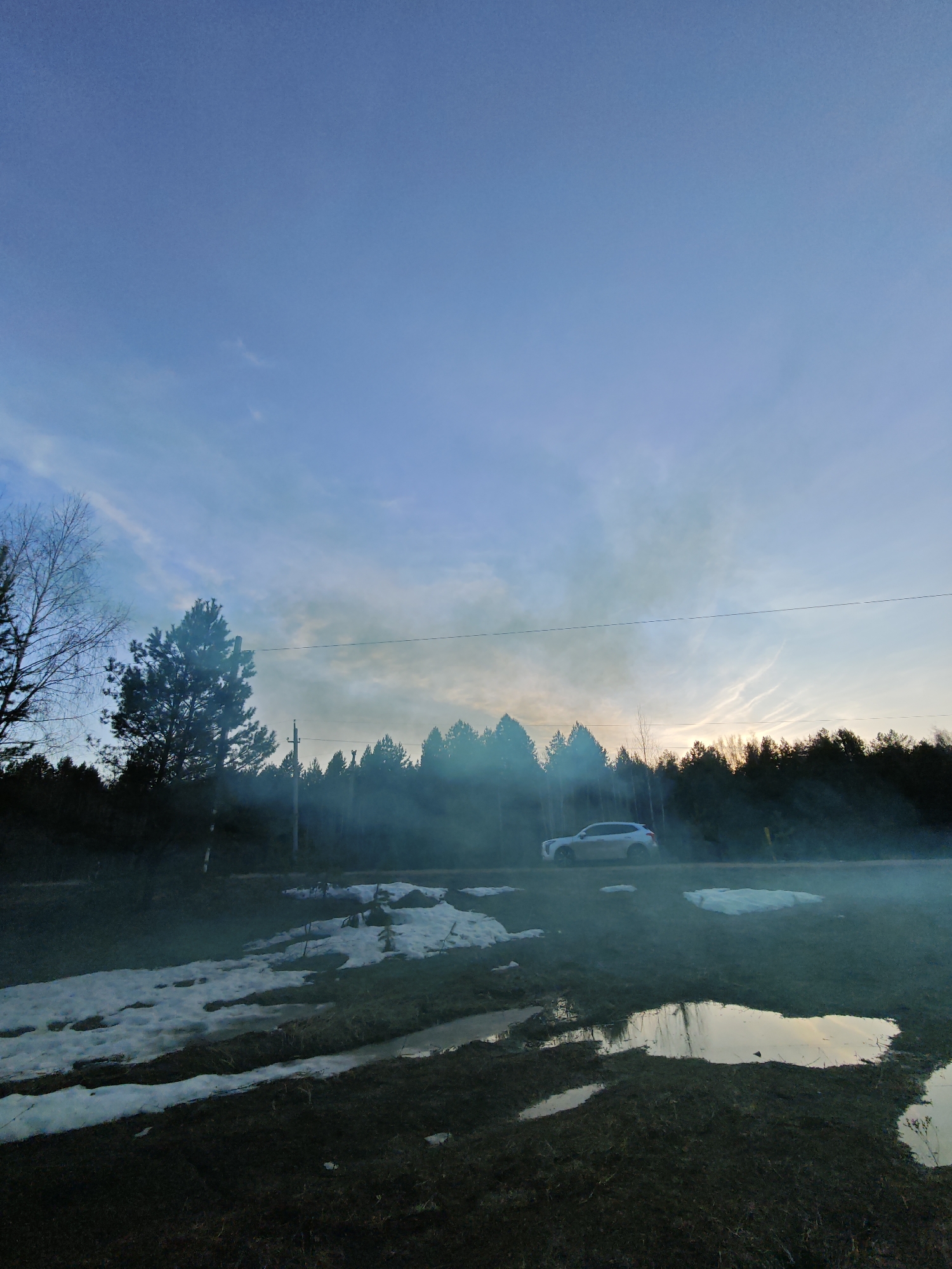 The image captures a rural landscape in Dubovaya, Russia, likely during either sunrise or sunset, given the warm light on the horizon and the cool blue of the sky above. The scene suggests a transition from late winter to early spring, with patches of melting snow on dark, damp ground.
A wide expanse of clear blue sky dominates the upper frame, subtly lightening towards the horizon. Faint, wispy clouds are visible, catching the soft light of the low sun. A dense line of dark coniferous trees forms the horizon line, silhouetted against the brighter sky, with a hint of warm light breaking through. To the left, a bare deciduous tree with intricate branches stands out.
The foreground features dark, muddy ground interspersed with irregular patches of snow. Several puddles reflect the pale sky, indicating recent melting. A significant amount of bluish-white haze or smoke permeates the air, particularly in the lower and mid-sections, partially obscuring the trees and creating a diffused atmosphere.
On the right side, a light-colored SUV or crossover vehicle, possibly white or silver, is parked on what appears to be a dirt road or shoulder, partially enveloped by the haze. Utility poles and a single power line are faintly visible stretching across the mid-ground. No visible text is present in the image. The overall impression is one of quiet transition in a natural, forested setting with lingering winter elements.