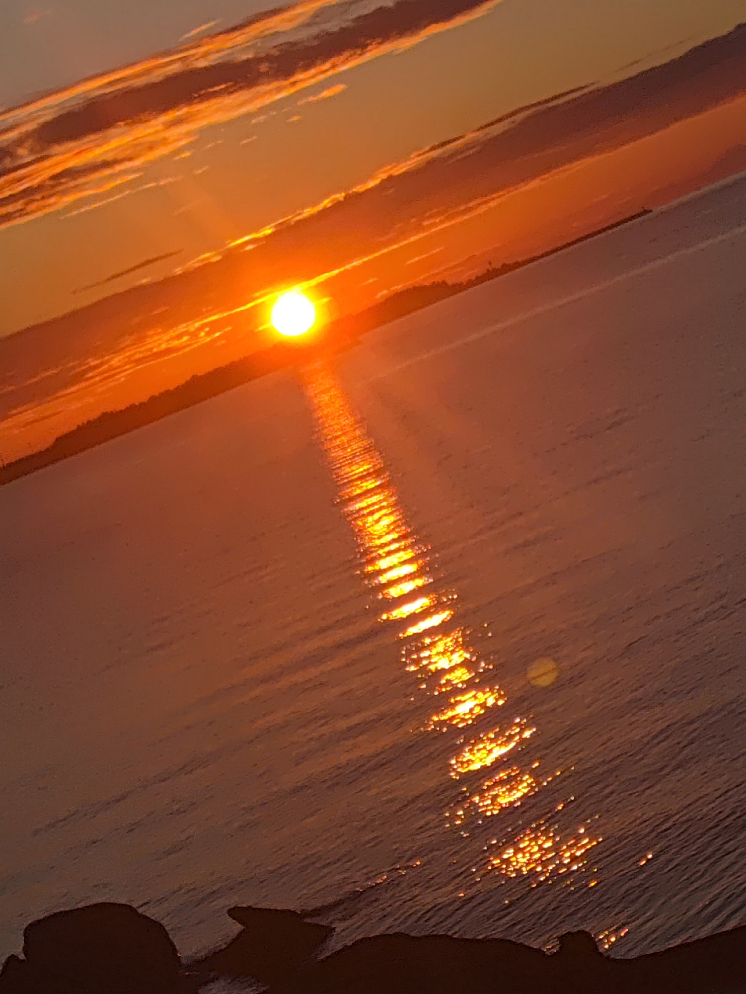 The image captures a sunset over the Baltic Sea, likely near Tallinn, Estonia, given the angle of the sun and the characteristics of the water and shoreline. The sun, a bright, glowing orb, is low on the horizon, casting a vibrant orange and yellow light across the sky and water. Streaks of clouds, illuminated by the setting sun, stretch across the upper portion of the frame, adding depth and texture to the scene. A shimmering reflection of the sun dances on the rippling surface of the water, creating a luminous path from the horizon to the foreground. The shoreline, visible in silhouette against the bright sky, appears to be lined with dark, rocky formations. The overall mood is one of serene beauty and natural spectacle, with the warm hues of the sunset creating a tranquil and picturesque panorama.
