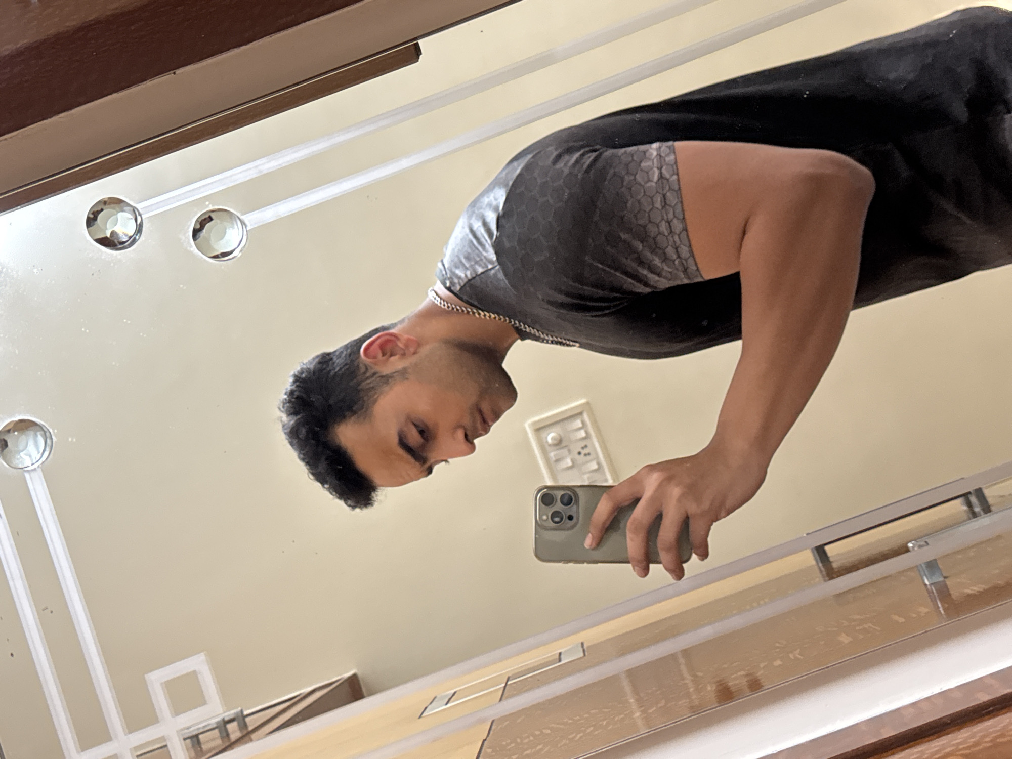 A person with short black hair is reflected in a mirror, holding a smartphone in their right hand. The person is wearing a black T-shirt with a gray patterned shoulder. A silver chain is visible around their neck. The background reflection includes a light beige wall with a light switch, and elements of a room with wooden furniture. The mirror has decorative circular accents and a multi-layered frame. Given the prevalence of such interior designs, the location could be a residential or commercial interior, but without specific identifiers, pinpointing the location to Kolkata, India, is impossible.