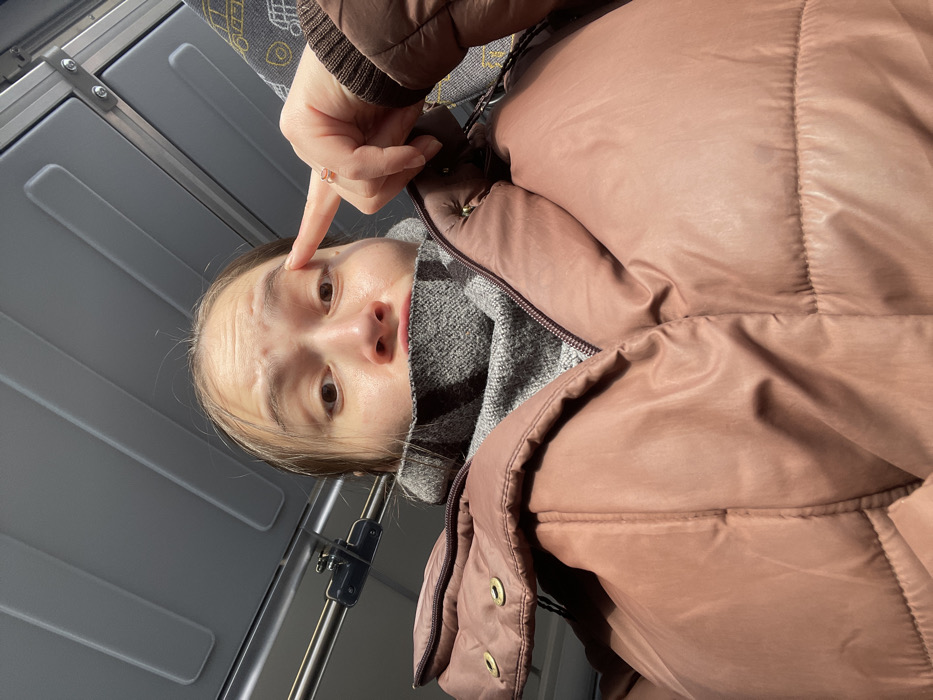 The scene depicts the interior of a public transport vehicle in Fryazevo, Russia. A female individual is centered in a low-angle, close-up shot, looking directly at the camera with wide-open eyes and slightly parted lips. She is dressed in a dusty rose-colored puffer jacket over a grey, textured high-neck garment. Her left hand is raised, with the index finger pointing towards her left eye; a ring is visible on her left ring finger. The background consists of a light grey paneled ceiling, a horizontal metal grab rail, and a segment of patterned upholstery, likely a seat.