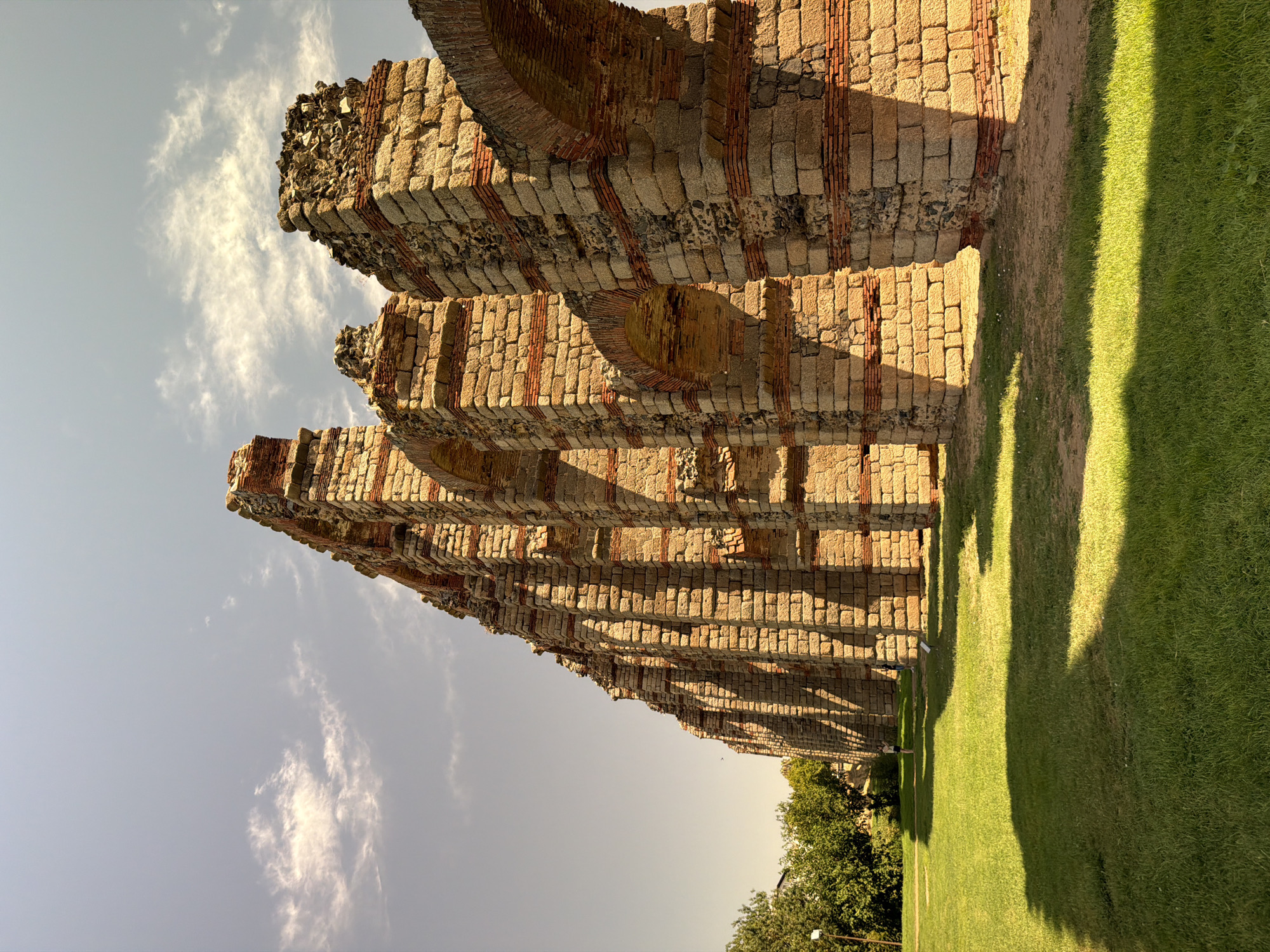 The image shows a section of the Roman Aqueduct of the Miracles in Merida, Spain, under a partly cloudy sky during daylight. The aqueduct is constructed of stone blocks and brick, featuring multiple arches, with some sections appearing weathered and partially collapsed. The aqueduct runs diagonally from the left foreground towards the right background, casting long shadows across a well-maintained grassy lawn. In the background on the left, there are trees and a hint of buildings. Two people are visible in the distance on the grassy area, one standing and looking towards the aqueduct, and another further back. The overall impression is of an ancient, grand structure set against a natural, open environment.
