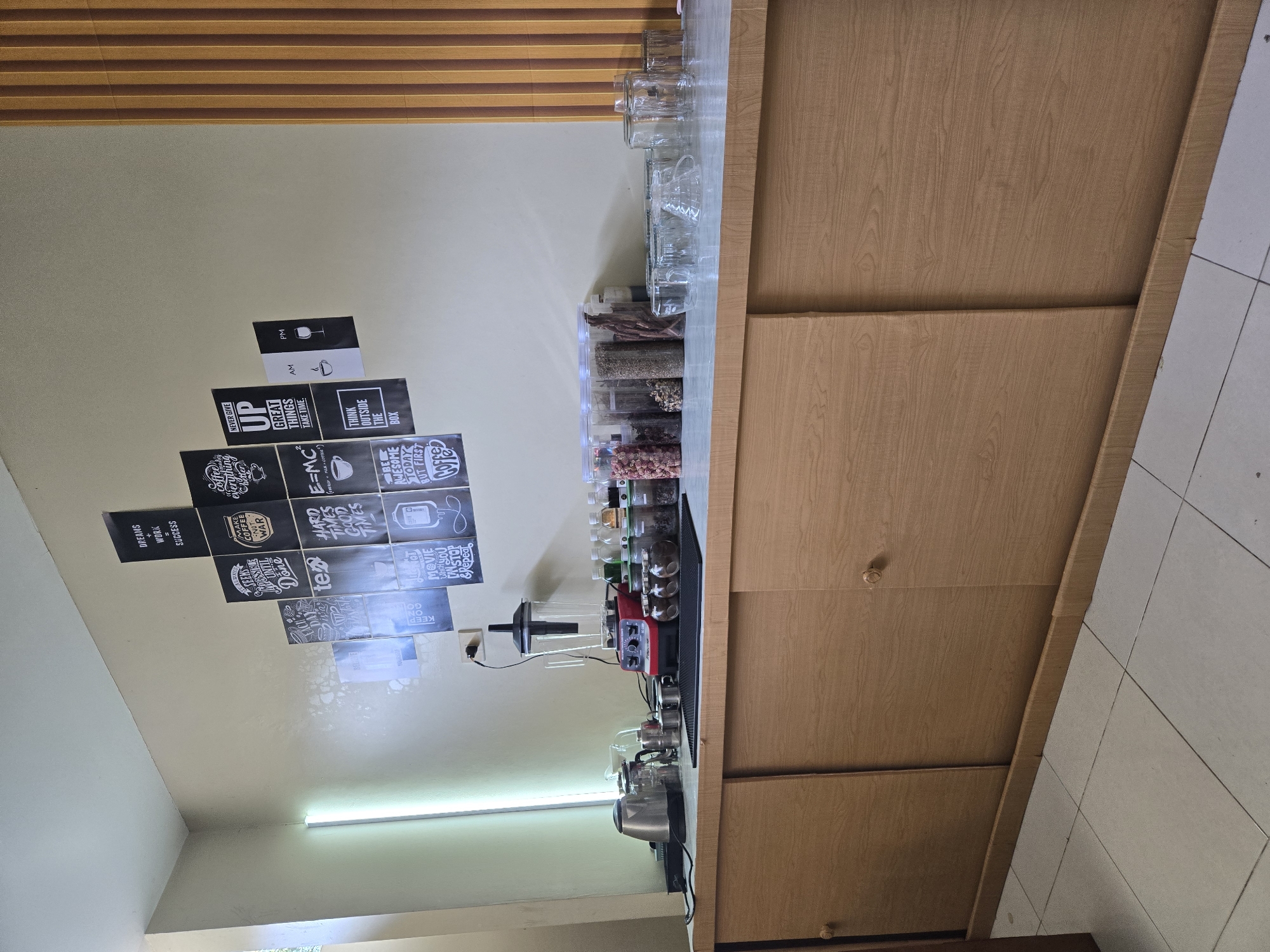 The image shows a coffee station setup in what appears to be a café or a dedicated coffee corner in a room in Sungailiat, Indonesia. The wall behind the counter is decorated with various motivational posters in black and white, featuring quotes related to coffee, dreams, success, and thinking outside the box.
On the counter, a red and black blender is positioned centrally. To its left are kitchen appliances including a silver electric kettle and a smaller silver teapot with a strainer. To the right of the blender are several clear glass jars filled with coffee beans and other granular ingredients. Further to the right, there are stacks of clear glasses of various sizes. The counter itself is a light-colored countertop, and it sits atop a wooden cabinet with sliding doors. The cabinet has a light wood grain finish and simple circular wooden handles.
The floor is tiled with light-colored square tiles. To the left of the counter, a fluorescent light fixture is visible on the wall, casting a soft glow. The overall scene suggests a functional and aesthetically pleasing coffee preparation area.