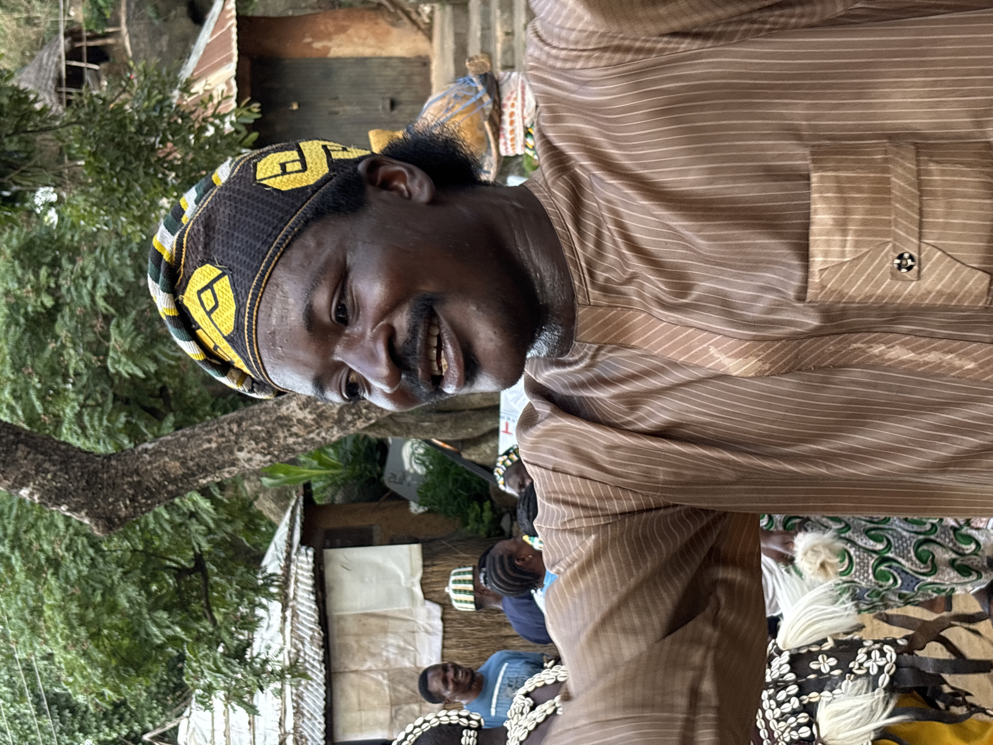A close-up, waist-up, medium shot shows a smiling Black man in the foreground wearing a brown, pinstriped kaftan and a black, yellow, and green cap. He is looking to his right and appears to be speaking or laughing. The background is a village scene in Jos, Nigeria, with thatched-roof buildings and lush greenery. Other people are visible in the background, some wearing traditional clothing, suggesting a cultural event or gathering. The lighting is natural, indicating it was taken outdoors during the daytime.