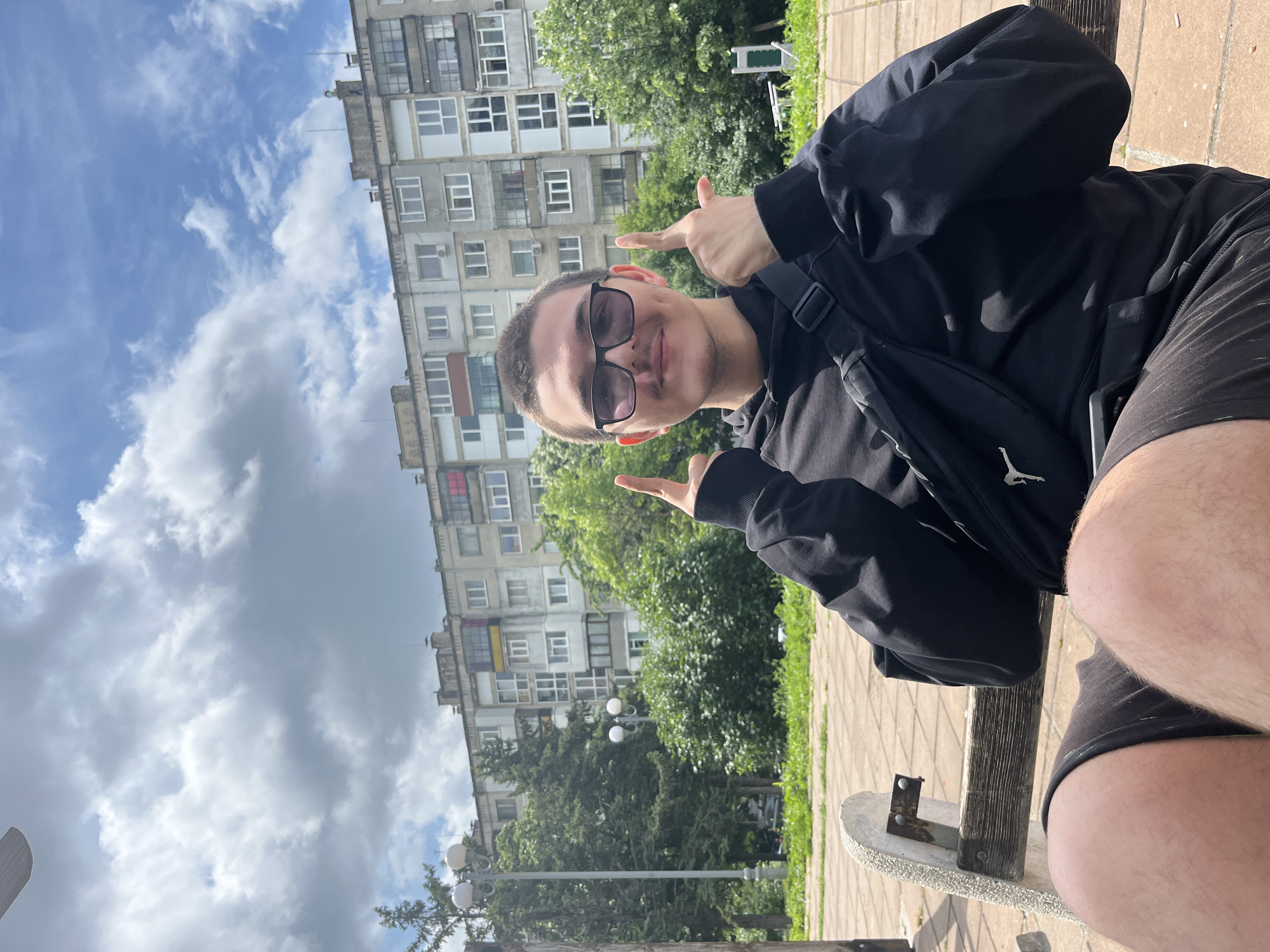 The image, correctly oriented in portrait format, captures a young man seated outdoors in Shumen, Bulgaria, looking directly at the camera with a playful expression. He is smiling and making a "finger gun" gesture with both hands, pointing to the sides. He wears dark sunglasses, a black hooded jacket, and black shorts, with part of his right leg visible. A black shoulder bag with a white Jumpman (Jordan) logo is worn across his body.
The background features a multi-story, concrete apartment building with numerous windows and balconies, typical of Eastern European residential architecture. Lush green trees and bushes are visible around the building and in the mid-ground. The man is sitting on a concrete and wooden bench, with a paved path or ground made of square stones visible in the foreground. The sky above is blue with scattered white clouds, indicating a bright and sunny daytime, likely late morning or early afternoon. The overall scene suggests a relaxed moment in an urban park or residential area.