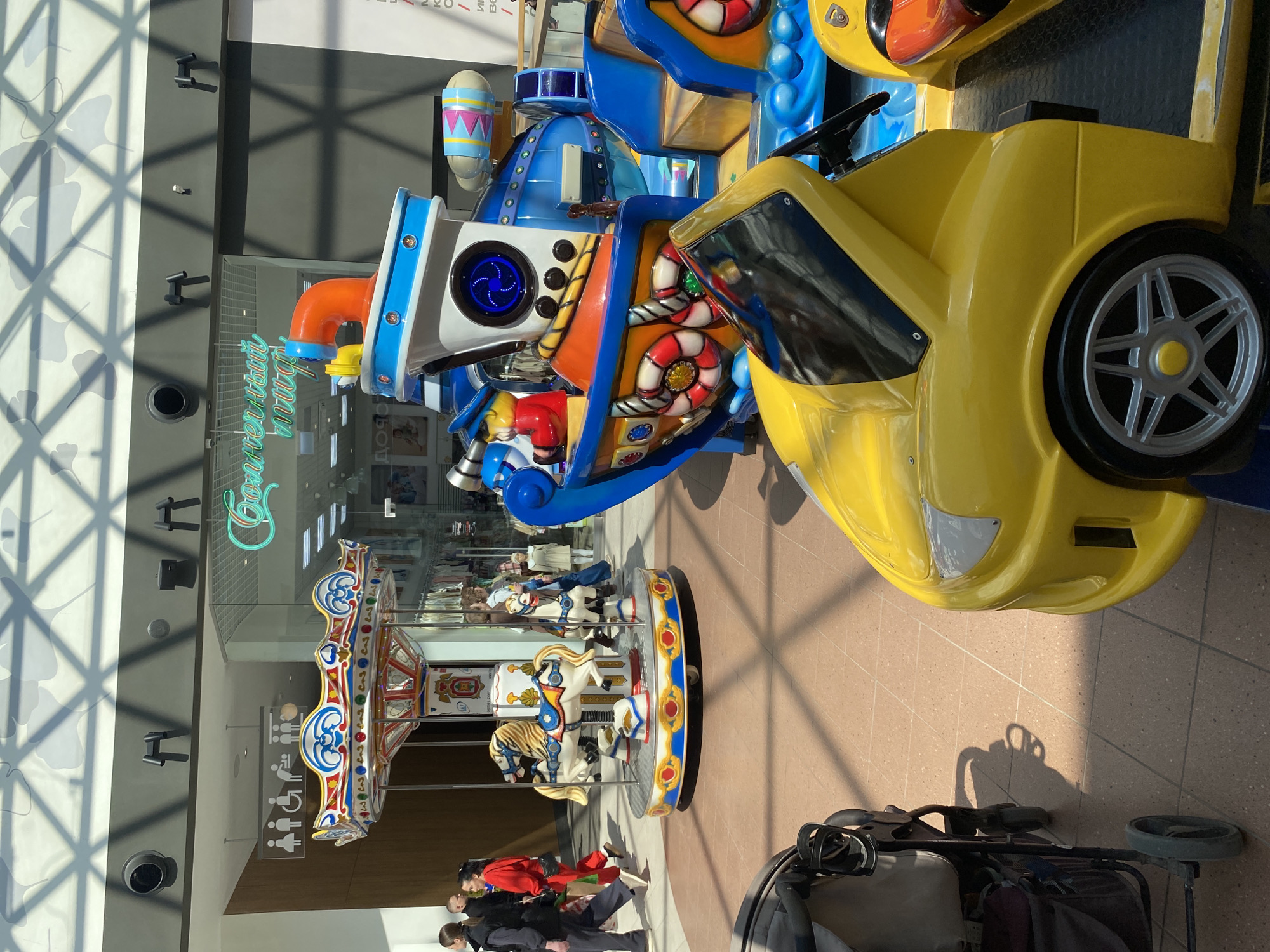 The image depicts an indoor scene, likely a shopping mall or entertainment center in Perm, Russia.
Dominating the foreground are several coin-operated ride-on toys, including a blue and white nautical-themed ride and a yellow car-shaped ride. Behind them, a small carousel with white horses is visible. In front of the carousel, three people are standing and interacting, partially obscured by the carousel base. A stroller is also in the foreground, further down.
The backdrop features the entrance to a store with a neon sign (likely in Russian). The architectural elements of the building include glass panels and metal frameworks, casting shadows on the walls. The floor is tiled.
Overall, the scene suggests a children's entertainment area inside a commercial establishment.