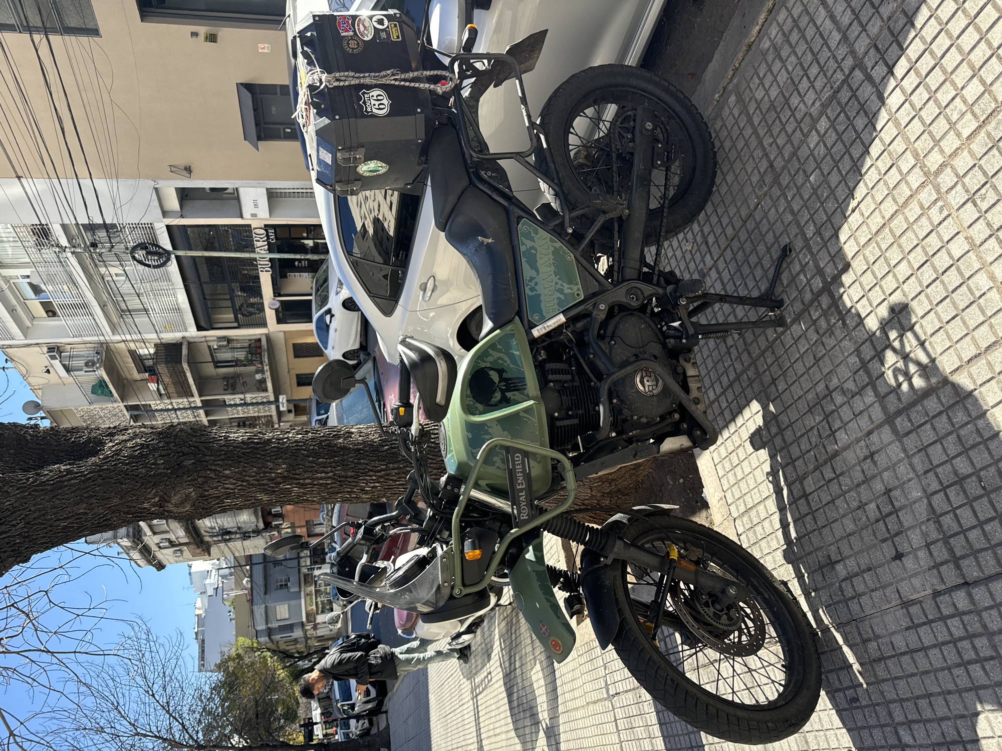 A Royal Enfield motorcycle with saddlebags is parked on a gray tiled sidewalk in a residential area. The motorcycle is a utilitarian olive green color. A person is walking past the front of the motorcycle toward the left side of the frame. A car is parked next to the motorcycle. A large tree trunk is to the left of the motorcycle. Buildings are visible across the street with the name "BUCARO CAFE" written on one. The scene is bathed in bright sunlight. The location is Buenos Aires, Argentina.