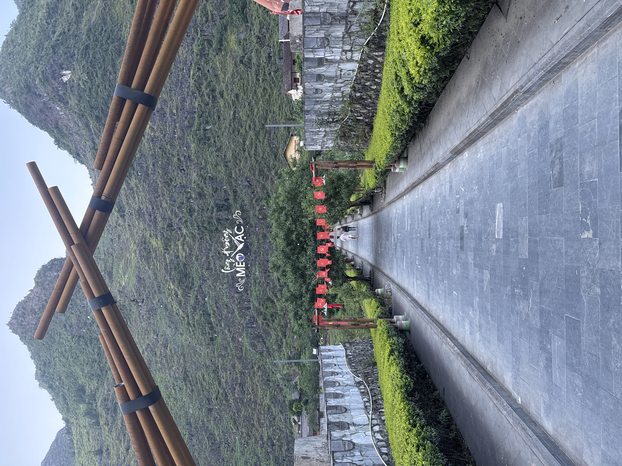 A landscape shot from Mèo Vạc Commune, Vietnam, featuring a stone path lined with hedges on both sides, leading away from the viewer. The path is composed of dark grey rectangular stones. Red flags are suspended above the path between wooden posts. Further back, the landscape is dominated by lush green mountains. A bamboo structure, resembling a large 'X', stands in the foreground. The sky is overcast, providing soft, diffused lighting. Two people are visible on the path, walking away from the camera. A structure with a stonework facade is adjacent to the path. A sign with "Mèo Vạc" is visible in the background.