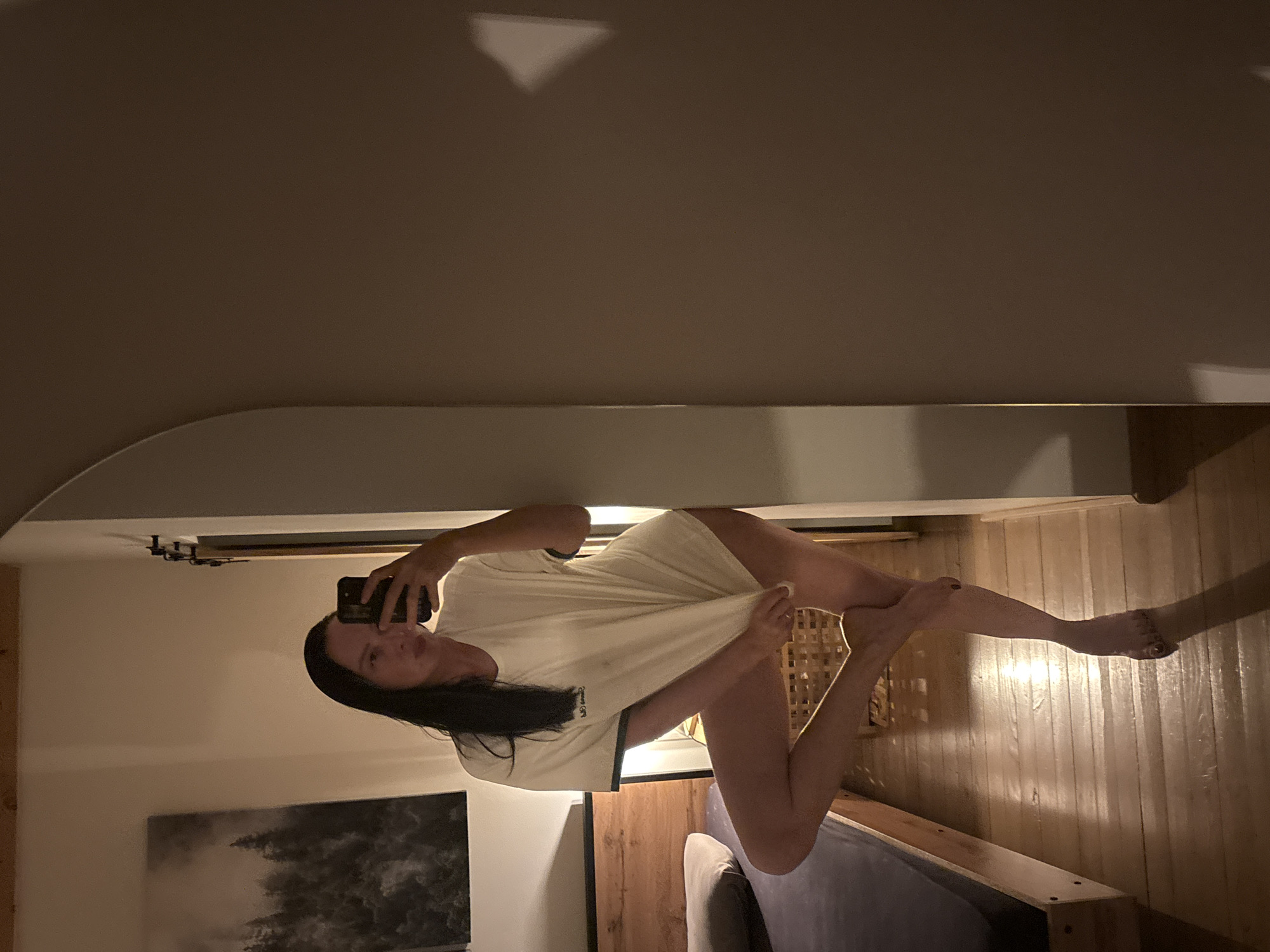 A woman takes a photo of herself in the mirror, holding her phone in her left hand. She is wearing a loose, light-colored T-shirt. The room contains a bed, a piece of artwork on the wall, and wooden paneling. The environment is dimly lit. The location is unknown.