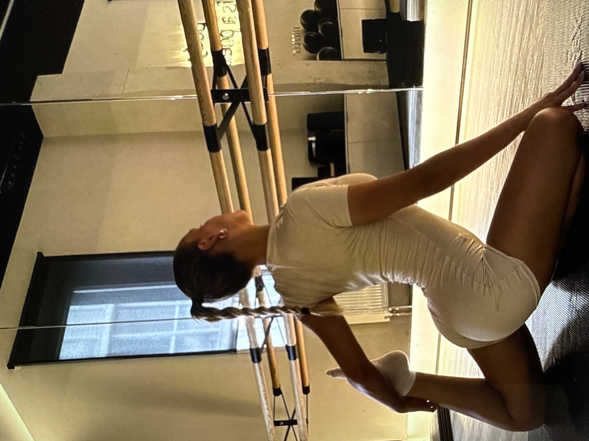 A woman in a white, short bodysuit and white socks kneels on a black, textured surface. She is stretching, leaning back with her arms reaching towards the floor. Her hair is braided and pulled back in a ponytail. She is in a dance or exercise studio, with ballet barres along the wall and a mirror behind her. A window with a dark shade is visible in the background. The location cannot be determined to be Sofia, Bulgaria from the image itself.
