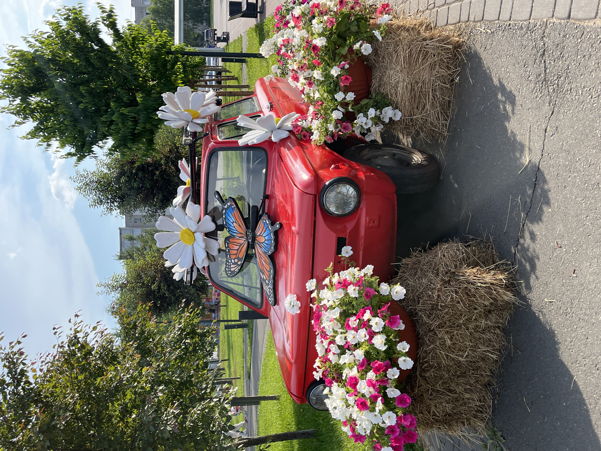 A vintage red car, possibly a ZAZ 968M Zaporozhets, is decorated with large white daisies and a butterfly decal. The car is parked on an asphalt surface, next to a cobblestone path. Two hay bales are placed on either side of the car, acting as stands for large planters overflowing with pink and white petunias. In the background, there are trees, a grassy area with some playground equipment, and apartment buildings, suggesting an urban park or public space. The sky is partly cloudy, and the sun is shining, casting shadows on the ground. There are no people clearly visible in the foreground, but there may be distant figures in the background. The overall scene appears to be an outdoor art installation or decorative display.