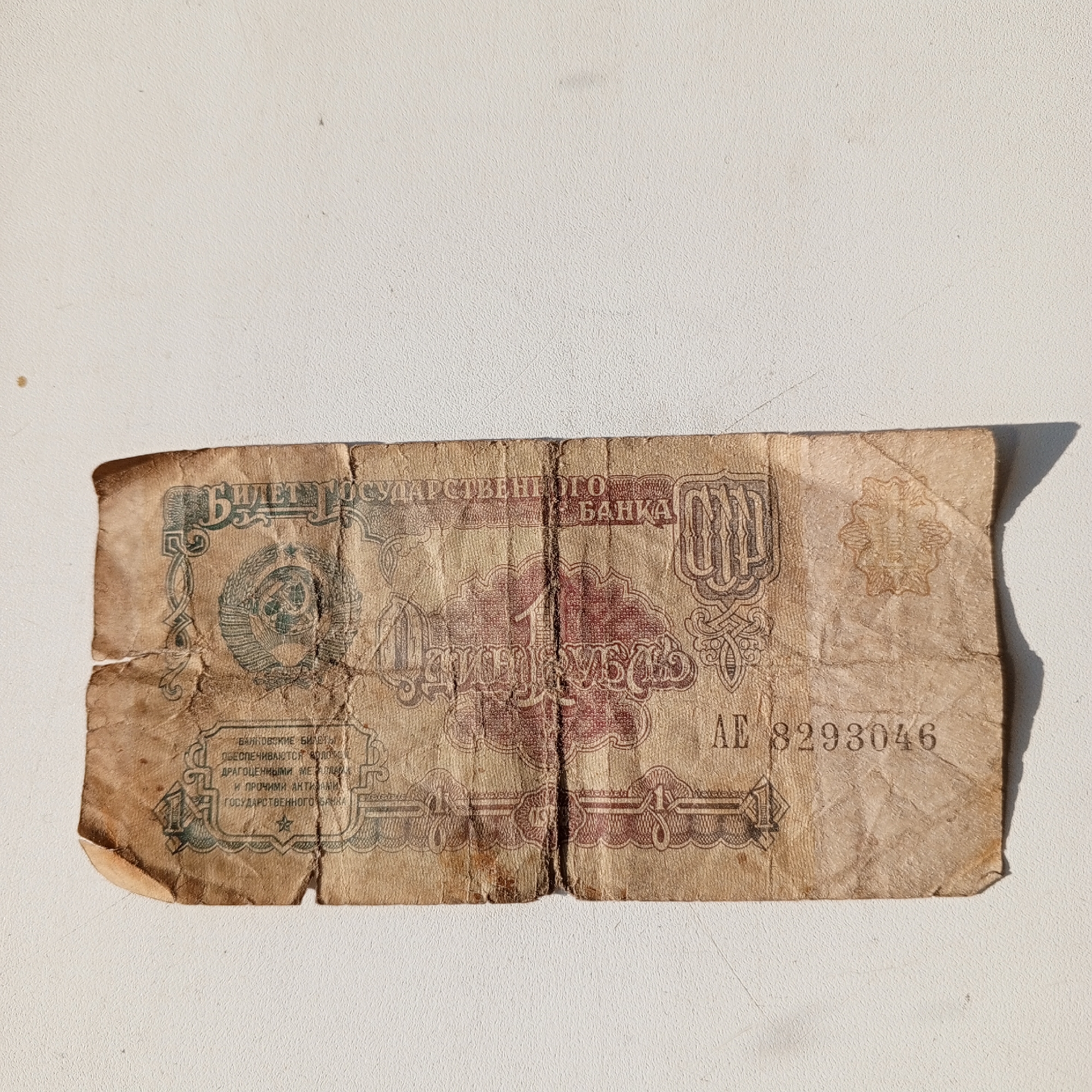 This is a close-up, eye-level, photograph of a crumpled, aged Soviet banknote lying on a textured, off-white surface, likely a table or floor. The banknote appears to be a 1 ruble note from the Soviet era, with Cyrillic text and an image of the hammer and sickle emblem. The paper is worn and creased, with some discoloration and visible folds. The lighting suggests it was taken indoors under natural light, casting a slight shadow to the right of the banknote. There are no people visible in the image, and the background is an indistinct, textured surface. The location in Sayanogorsk, Russia, is not visually identifiable from the image itself.