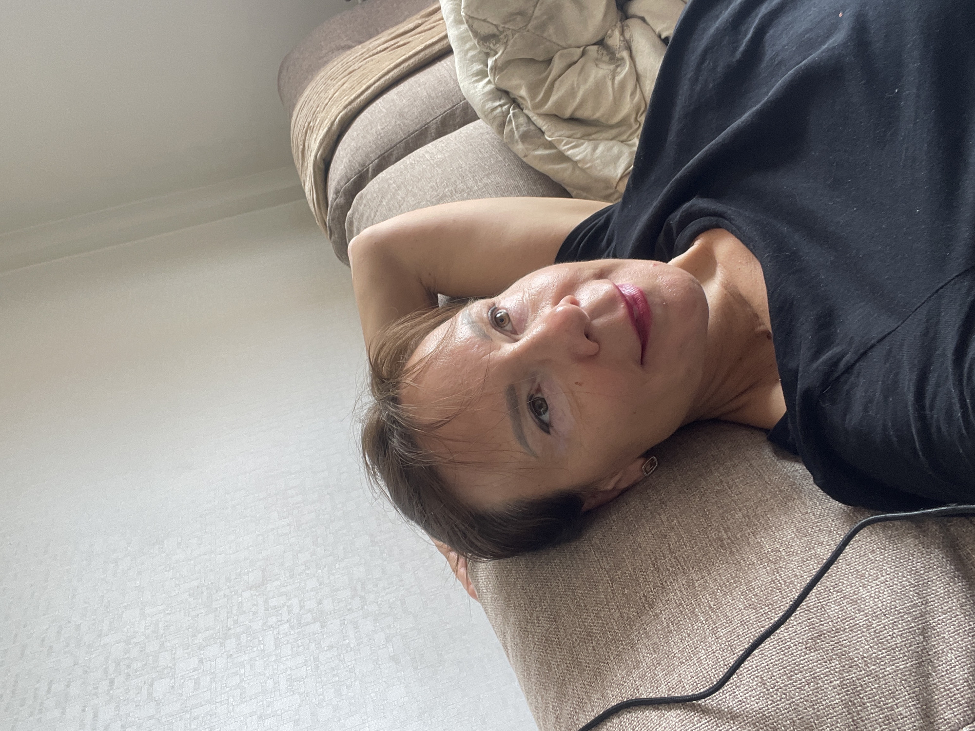 A woman is lying on a couch in a room. She has short brown hair and is wearing a black t-shirt. Her right arm is bent behind her head, and her left hand is resting on her chest. She is looking to the right and appears to be relaxed. The couch is covered in a beige fabric, and there is a folded blanket on the right side of the couch. The wall behind the couch has a textured pattern. The scene is indoors, and there is natural light coming from the right. The image does not contain any information to determine the location in Sochi, Russia.