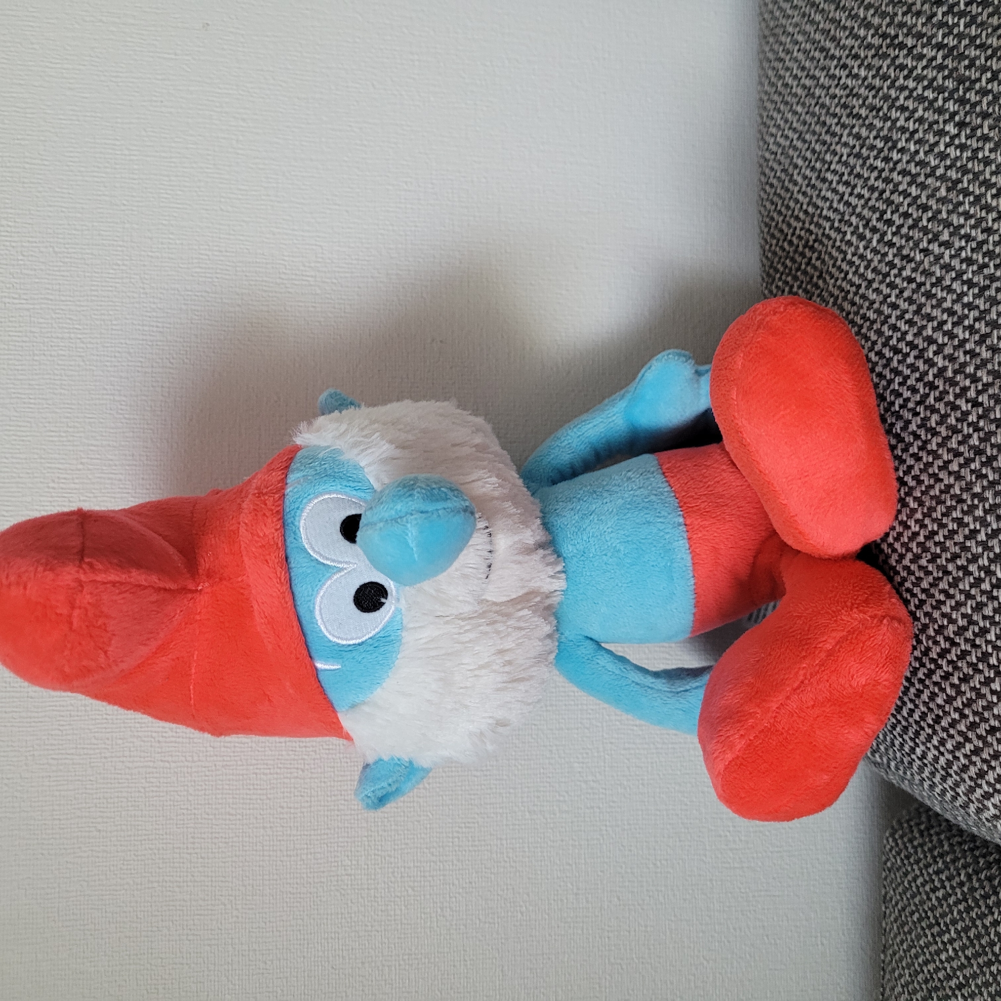 A plush toy Smurf is sitting on a patterned sofa against a white textured wall. The Smurf has a blue body, a red hat, red shorts, and white fur around its face. Its eyes are large and white with black pupils, and it has a blue nose and a smiling mouth. The sofa is upholstered in a dark gray and white woven fabric. The background is a plain white wall with a subtle vertical textured pattern. The lighting appears to be from a natural source, casting a slight shadow to the left of the Smurf. The scene is static and composed of a single inanimate object. The location is not discernable from the image and is not in Jūrmala, Latvia.