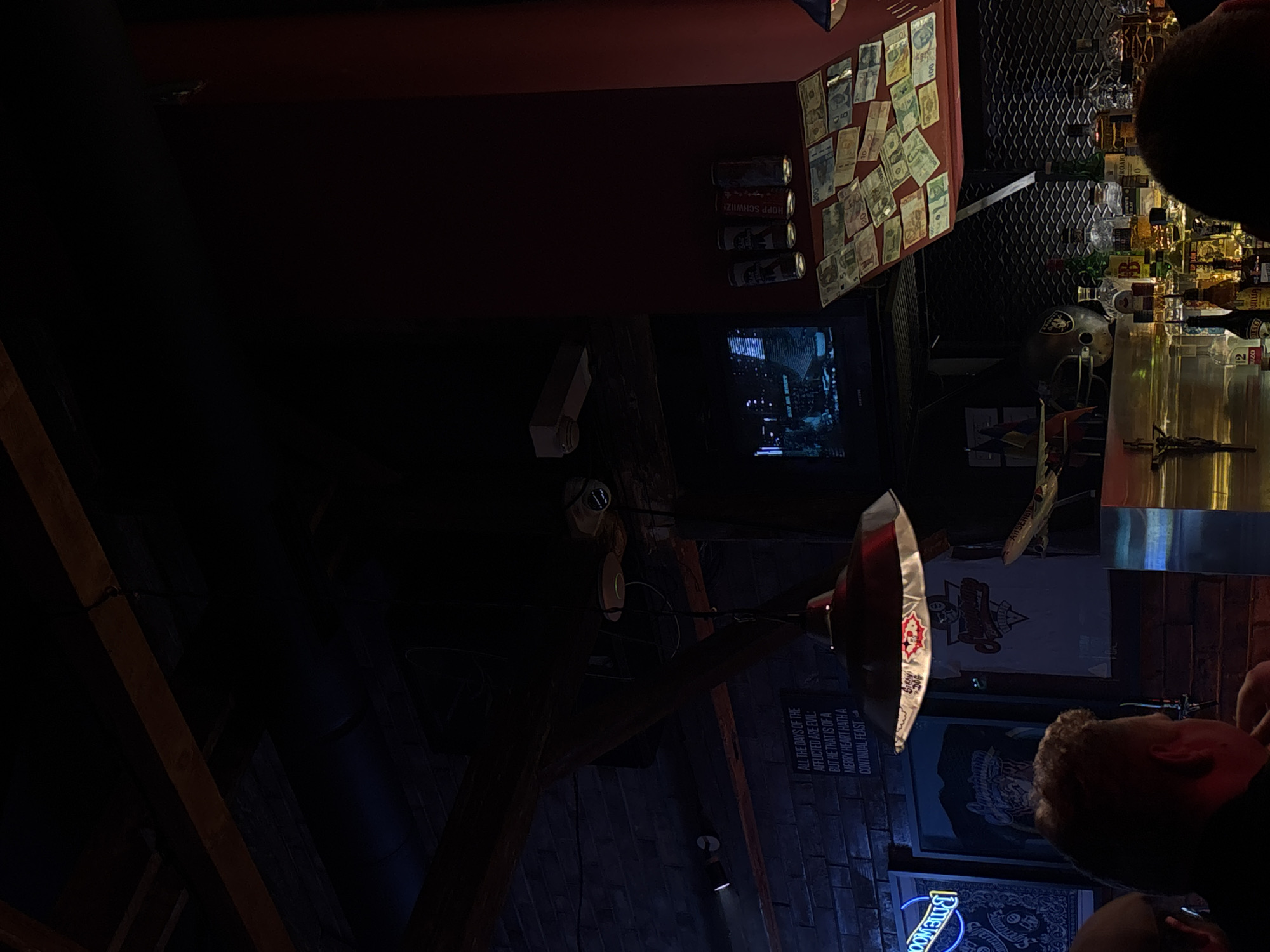 The image shows the interior of a bar in Belgrade, Serbia. The bar features a collection of foreign banknotes affixed to a red wall, beverage cans, and a television screen. Bottles are behind the counter and the bar's architecture is apparent with wooden beams. A sign on the wall reads "ALL THE DAYS OF THE AFFLICTED ARE EVIL: BUT HE THAT IS OF A MERRY HEART HATH A CONTINUAL FEAST." A man with fair hair is visible, and a portion of another person's head obscures the top of the frame.