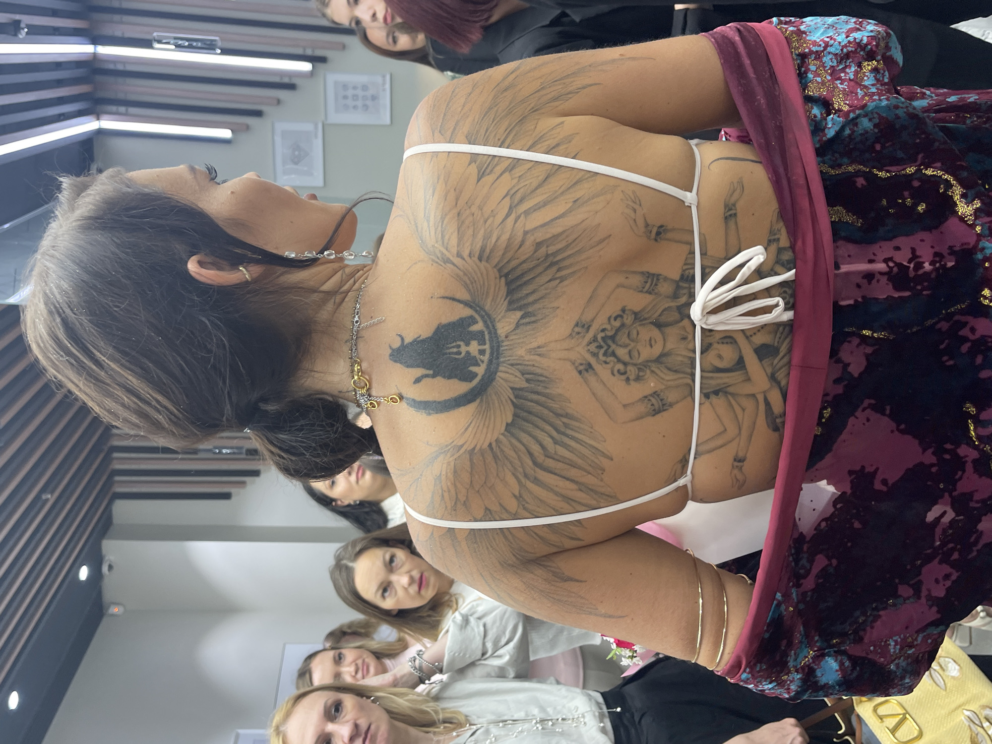 The image captures an indoor scene, likely an event or gathering, taking place in a modern commercial space in Moscow, Russia. The primary subject is a woman viewed from behind, turning slightly to her right, showcasing her left profile. She has dark hair styled in a ponytail and wears a white, backless halter-neck top. Her back is extensively tattooed with intricate designs.
The upper back tattoo features a silhouette of a figure, possibly a deity (suggesting Shiva with a trident), seated on a crescent moon, flanked by large, detailed feathered wings extending across her shoulder blades and upper arms. Below this, on her lower back, is another tattoo depicting a multi-armed female deity figure, adorned with a crown or elaborate headpiece, and positioned with multiple hands in various gestures.
The woman wears a gold chain necklace with a circular pendant and other small charms. A long, dangling earring with clear, tear-drop shaped elements is visible on her left ear, which also has a small gold cuff. Her left upper arm is adorned with three thin gold armbands. She has a loose, colorful garment, possibly a wrap or jacket, draped over her forearms and lower back, featuring a maroon and dark purple base with splashes of teal and gold glitter.
In the background, several women are visible, observing or attending the event. They are dressed in light-colored tops, including white, cream, and pale pink. The interior features light-colored walls and a dark ceiling with integrated horizontal light panels and recessed spotlights. A small security camera is mounted on the ceiling. On the walls, framed artwork or diagrams, one clearly showing a diamond shape, suggest a connection to jewelry or design. A yellow designer bag with a prominent "V" logo is partially visible on the floor to the lower left, and a small bouquet of pink and white flowers is partially visible behind the main subject's left side.