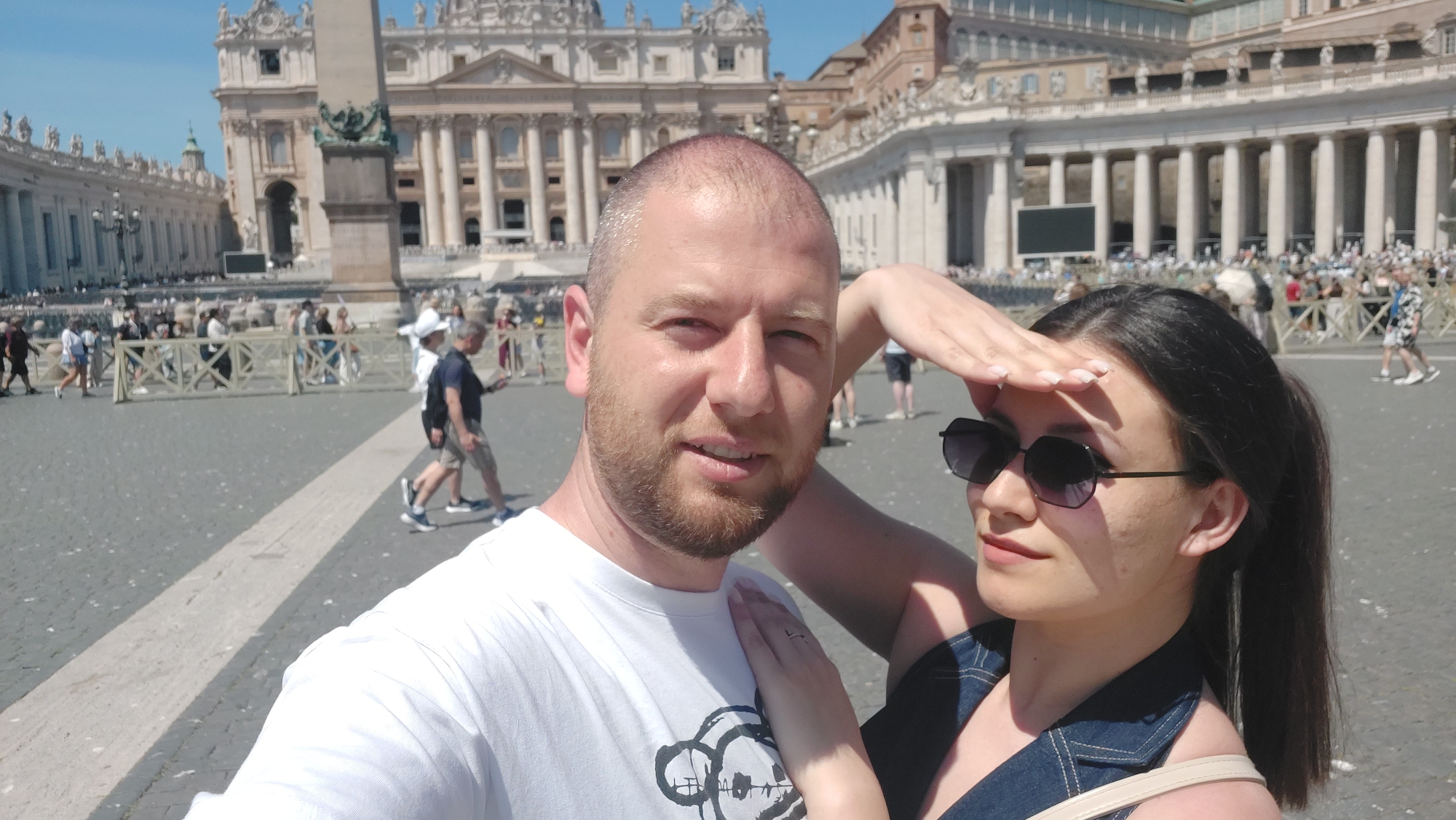 The image captures a selfie of a couple in St. Peter's Square, Vatican City, Roma, Italy. The man has a receding hairline and a beard, wearing a white t-shirt with a graphic design. The woman wears sunglasses and a denim top, shielding her eyes with her hand. Behind them, the square is bustling with tourists and pedestrians. In the background, the architecture of St. Peter's Basilica and the colonnades of the square are visible. The scene is bathed in bright daylight.