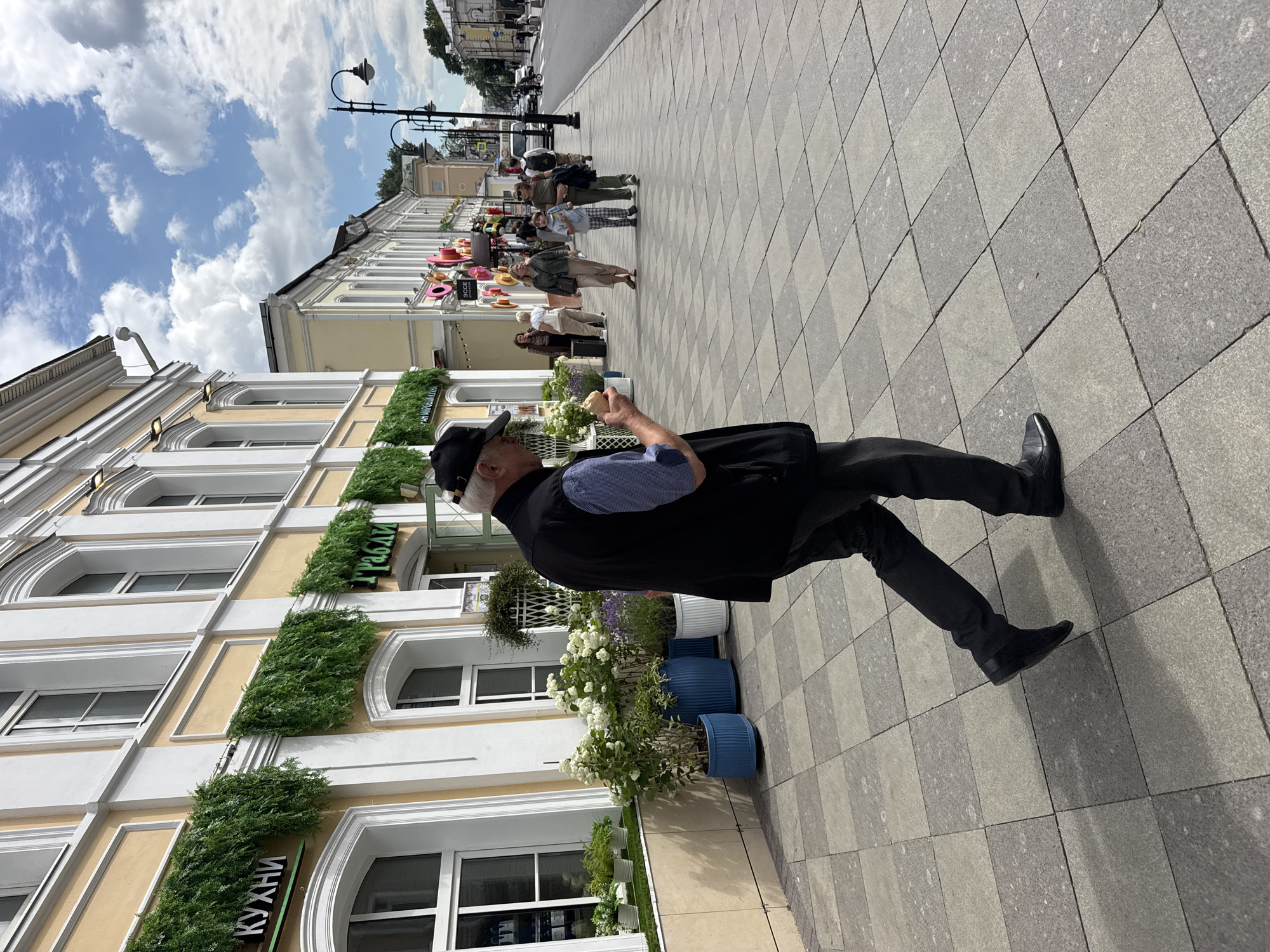 An elderly man wearing a black vest, blue checkered shirt, black pants, and a black baseball cap walks on a checkered sidewalk in Moscow, Russia, holding an ice cream cone in his right hand. The man is in the foreground, and his back is turned to the camera. He is walking on a sidewalk made of light gray and dark gray square tiles.
Behind him, on the left, is a light yellow building with white trim and arched windows. Green artificial plants hang from the facade of the building. A sign with Cyrillic text is visible above one of the windows. Several potted plants, including white flowering bushes and lavender, are arranged along the base of the building.
In the background, people are walking on the sidewalk. Further down the street, there are more buildings, lampposts, and a road with vehicles. The sky is blue with white clouds. The scene appears to be a street in a city on a sunny day.