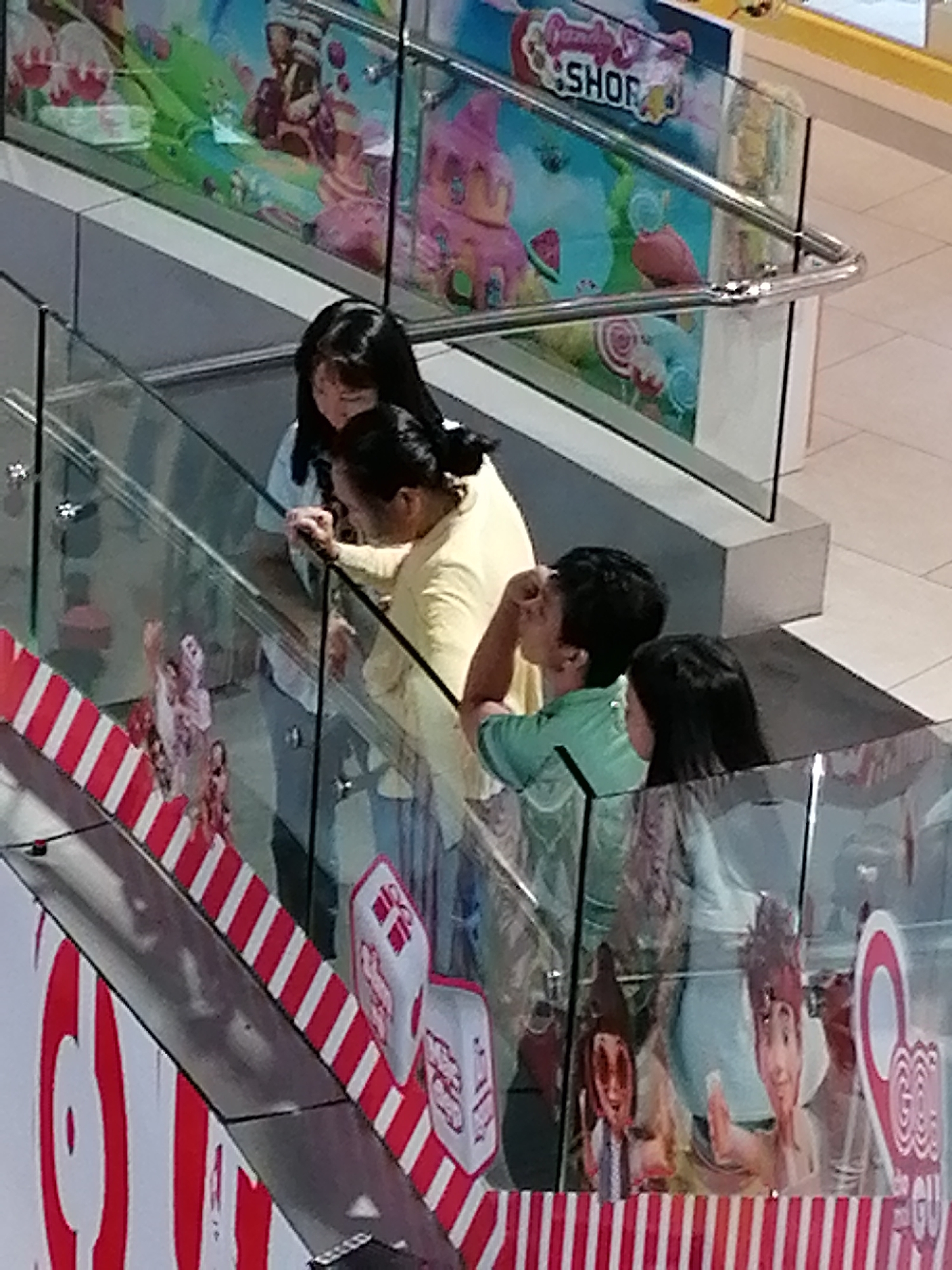 The image captures a group of individuals on an escalator, likely within a shopping mall. In the foreground, an escalator railing is visible with a red and white striped design. Behind the railing, two people are standing, one appearing to be a woman with long dark hair and the other a man with dark hair wearing a green shirt, leaning forward with his hand to his face. Further up the escalator, another woman with dark hair and a yellow top is visible, looking down. Above them, a colorful mural depicting candy and sweets adorns a wall, with the words "Candy Shop" prominently displayed. The overall scene suggests a commercial setting, possibly a shopping mall or entertainment venue in Da Lat, Vietnam.