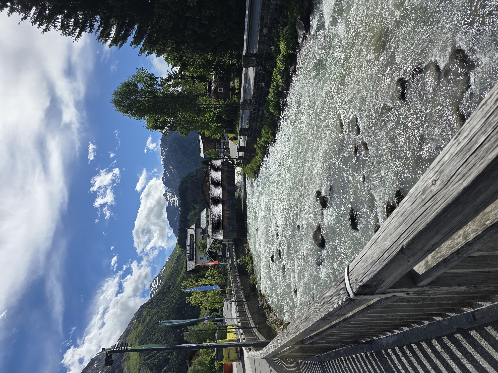 A wide shot captures a fast-flowing, rocky river in Lech, Austria. The river is in the foreground and center of the image, with a wooden fence on the left and a stone wall on the right. Beyond the river, a village with traditional wooden buildings and a church is nestled against a backdrop of green, tree-covered mountains. Snow-capped peaks are visible in the distance. A yellow bus is parked on the road, and a few people are walking or biking. The sky is blue with scattered white clouds. The scene appears to be a sunny day in a mountainous region.