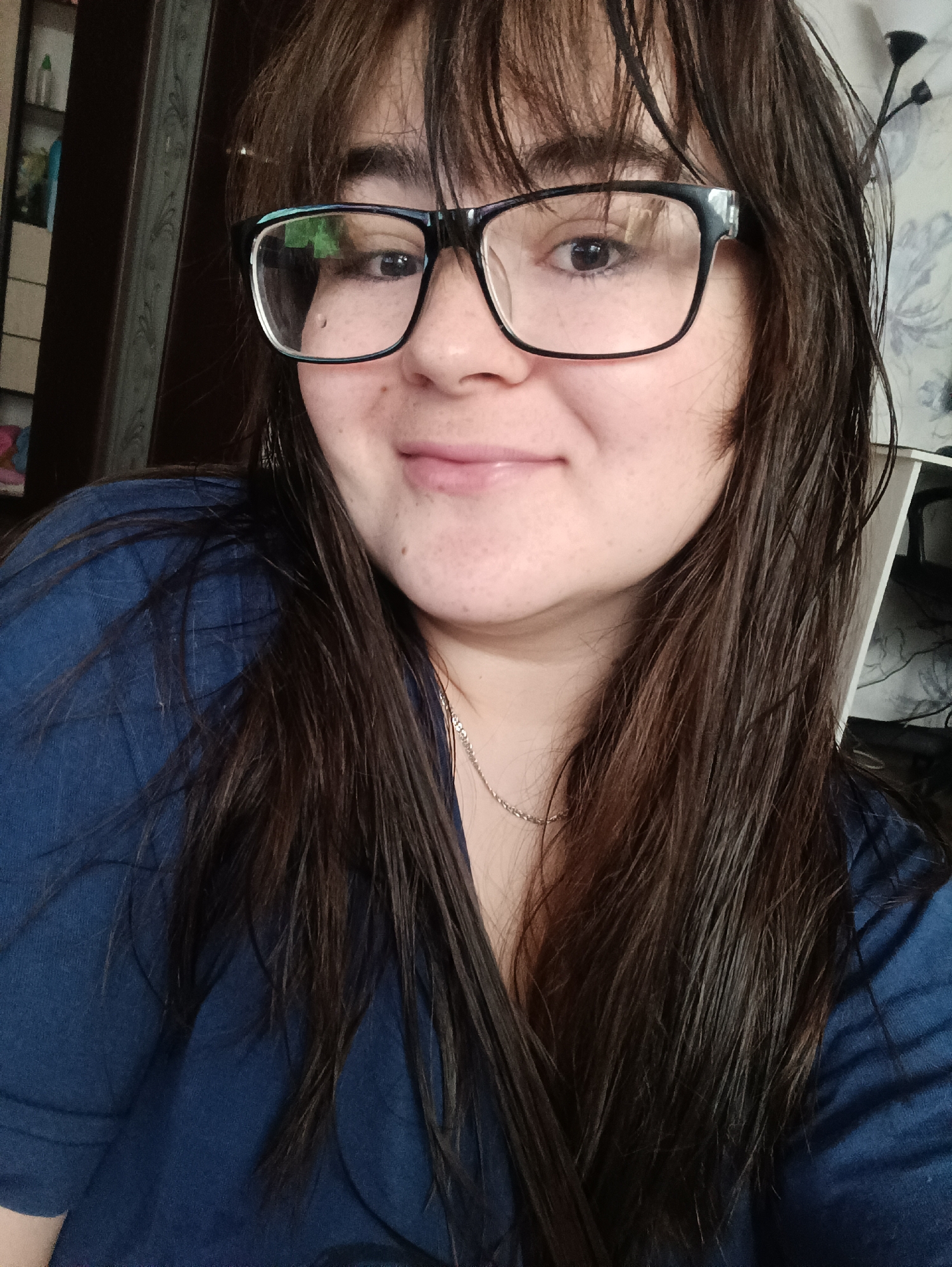 A close-up photograph shows a woman with dark brown, wet hair and bangs. She is wearing black-framed glasses. She has fair skin and is wearing a dark blue top. A thin silver necklace is visible around her neck. The background is partially visible and includes a dark brown door frame and a patterned wallpaper. The location, Noviy, Russia, cannot be verified from the image content.