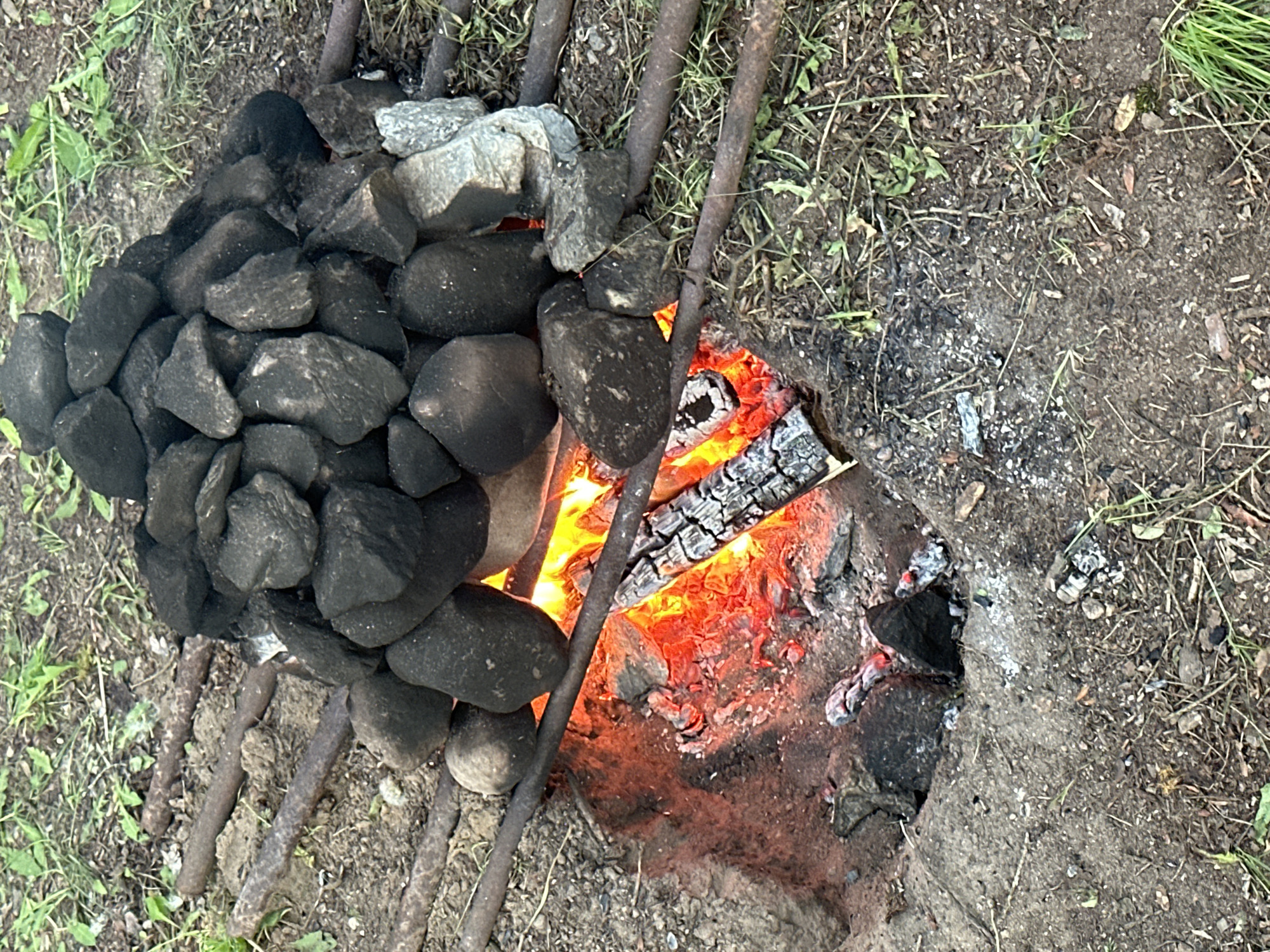 The image shows a fire pit located outdoors, likely in a rural area based on the surrounding grass and dirt. The fire pit is constructed in a hole in the ground. Flames are visible, burning wood inside the pit. On top of the pit is a metal grate, with a pile of dark, rounded stones arranged on the grate. The scene is lit by natural daylight. No people are visible. The image does not contain enough information to confirm the location as Папярнянскі сельскі Савет, Belarus.