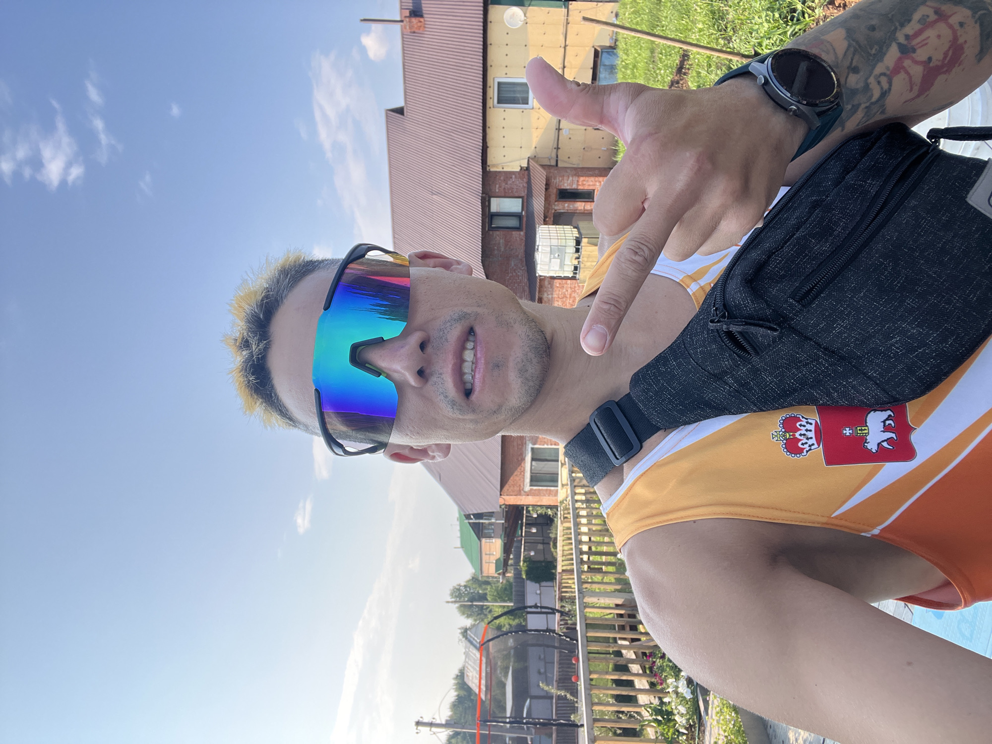 A man with dyed blond hair and rainbow-colored sunglasses is taking a selfie outdoors. He is wearing an orange and white tank top with a coat of arms emblem on it, and a black fanny pack is slung across his chest. He is giving a middle finger gesture with his right hand and his left hand is visible. In the background, there are houses, a wooden fence, a trampoline, and lush green vegetation under a clear blue sky. The scene appears to be a residential area in the city of Chaikovsky, Russia, possibly a dacha or suburban property.