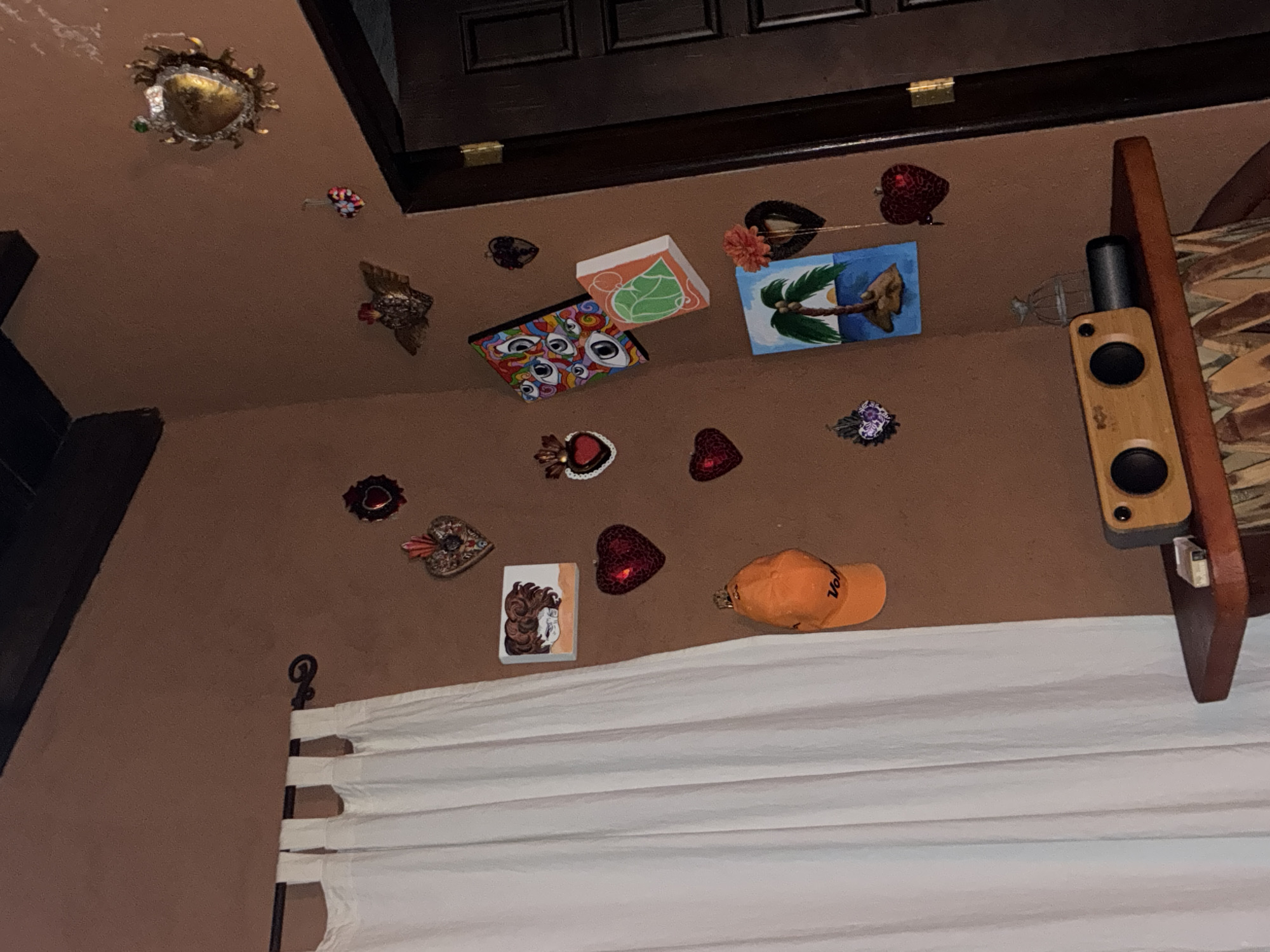 This image shows the interior of a room, likely in Cañada de Madero, Mexico, as suggested by the presence of cultural elements.
The room has a brown wall decorated with various items. Above a white curtain are several decorative objects, including hearts, artwork, and an orange baseball cap. On the right side of the wall, there is a wooden shelf with a speaker and what appears to be a bed with a patterned headboard.
The items on the wall have a colorful and folk art aesthetic. They include small paintings, possibly depicting local scenes or religious symbols, and other decorative pieces, suggesting a personal and culturally rich space.
The white curtain is slightly wrinkled. The room's lighting is subdued, contributing to the warm and inviting atmosphere.