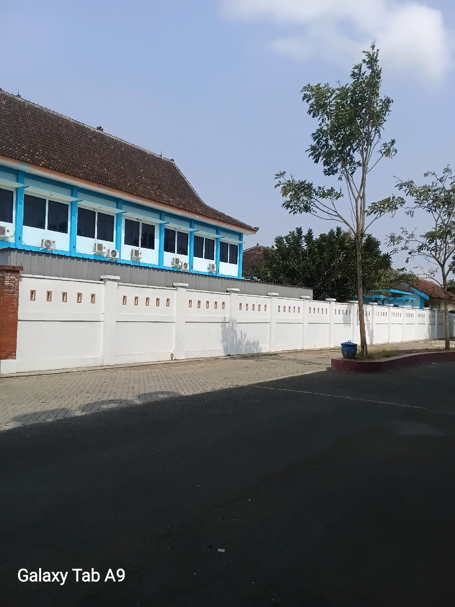 The image shows a long, white, concrete wall with square openings, topped with pillars. Behind the wall, a light blue building with a brown tiled roof and multiple windows is visible. Several air conditioning units are mounted beneath the windows. In front of the wall, a paved walkway runs alongside it, with a dark asphalt road to the right. A tall, slender tree with green foliage stands to the right of the wall, with a dark blue cylindrical planter at its base. The sky is clear and blue. The text "Galaxy Tab A9" is visible in the lower left corner. The image was likely taken in Trenggalek, Indonesia, given the watermark.