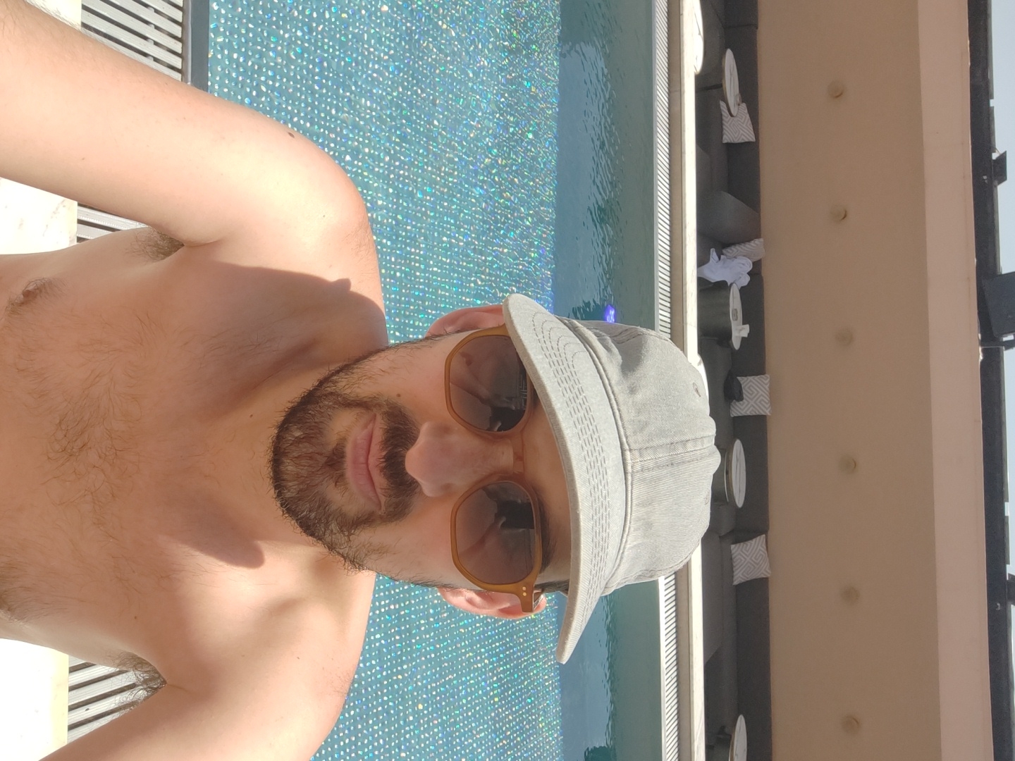 A fair-skinned man, seen from the chest up, is posing for a photo outdoors, likely a selfie. He has a beard and mustache, wears amber-tinted sunglasses, and a light grey cap. His upper body is bare, showing a hairy chest and arms. He is looking directly at the camera with a relaxed expression.
Behind the man is a sparkling blue swimming pool. To the right of the pool, a light-colored wall is visible, adorned with four circular, white fixtures and some white objects or cushions with geometric patterns. The bright light and strong shadows suggest a clear, sunny day, likely around midday. The setting appears to be a resort or hotel pool area. No visible text is present in the image. The location is Tel-Aviv, Israel.