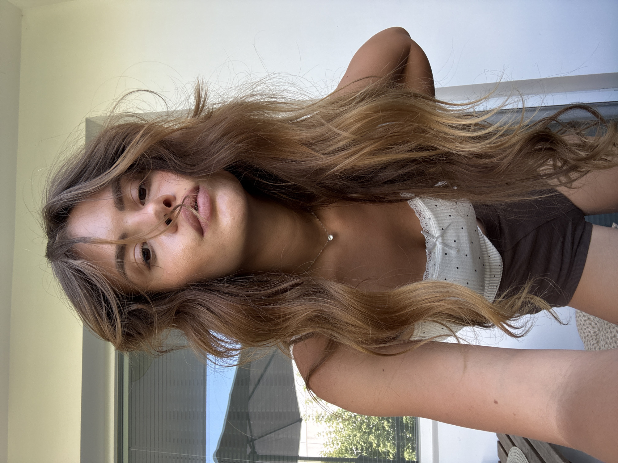 In Plovdiv, Bulgaria, a young woman with long, wavy brown hair poses for a selfie outdoors. She is wearing a white tank top with polka dots and a lace trim, and brown shorts. She has a delicate necklace with a single diamond pendant. The background shows a white wall and a window with blinds. A table and chairs are visible to the left, suggesting an outdoor seating area. The lighting indicates it is daytime, with sunlight casting soft shadows.
