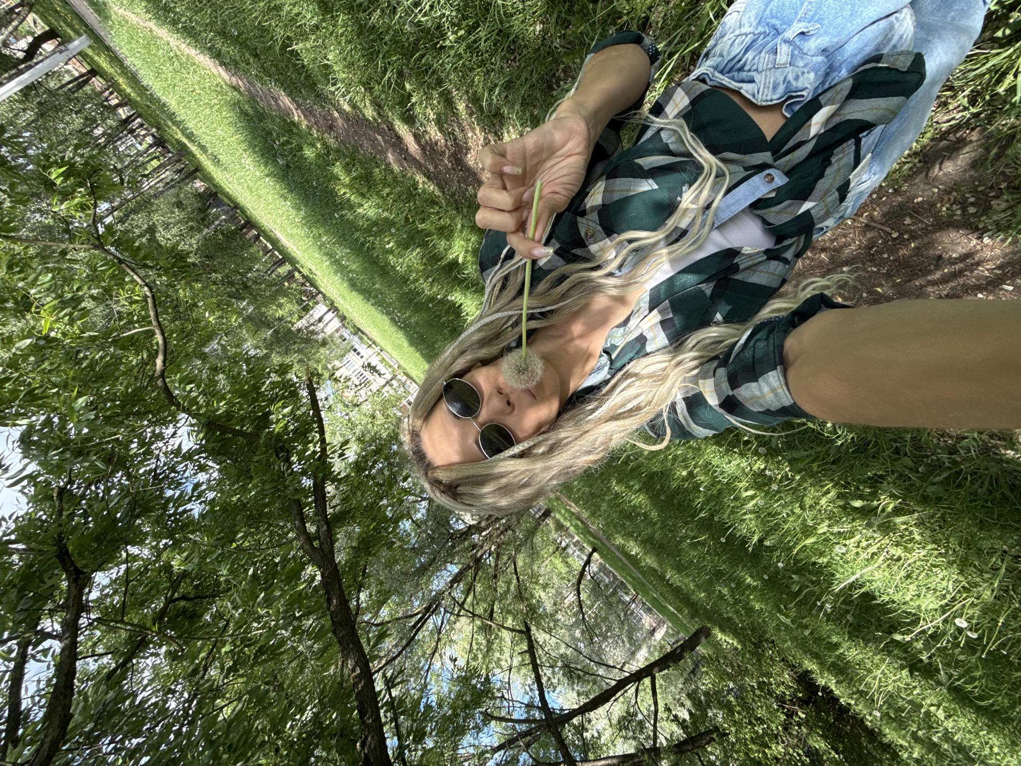 A woman is holding a dandelion in an outdoor setting with trees and grass. She is wearing sunglasses, a green and white plaid shirt, and blue jeans. The location appears to be a park or grassy area. The location is not specified to be Rusanovo, Russia.