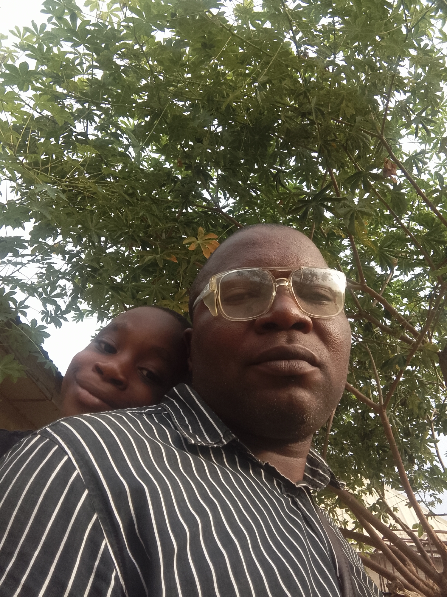 A close-up outdoor selfie captures an adult male and a child posing together. The adult male, with dark skin and prominent clear-framed glasses, is featured predominantly in the right foreground, looking directly ahead with a neutral expression. He wears a black shirt with narrow white vertical stripes. Behind him, on the left, a child with dark skin is partially visible, resting their head near the adult's shoulder. The child has a soft, slight smile on their face, looking towards the camera.
The background is dominated by the dense, vibrant green foliage of a tree, characterized by large, lobed leaves resembling those of a cassava plant. Bright, diffused daylight is visible through the gaps in the leaves, indicating clear weather during the daytime. The perspective suggests the photo was taken from below the tree, looking upwards. The overall mood is intimate and warm, highlighting the connection between the two individuals. There is no visible text in the image.