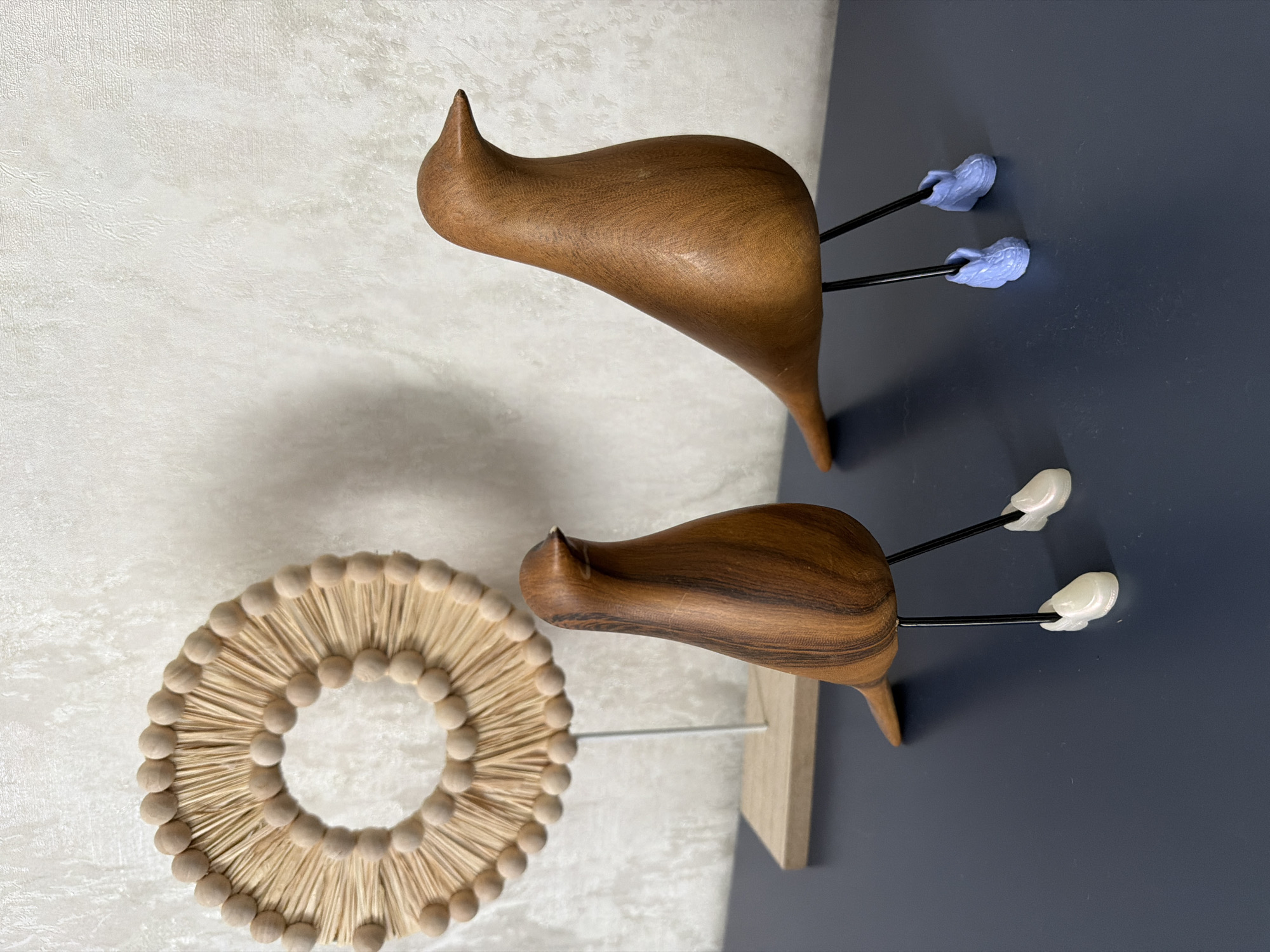 Two wooden bird figurines are positioned on a dark blue surface against a textured off-white wall. The bird on the left is smaller and closer to the viewer, its body displaying dark wood grain patterns. It stands on black wire legs with small white boot-like feet. To the left of the birds, a circular decorative object made of straw-like material and small wooden beads is mounted on a thin metal rod, which is supported by a light-colored wooden base. The larger bird on the right is further away, with a smoother, lighter brown wood finish. Its black wire legs are adorned with light blue, textured boot-like feet. The scene appears to be an indoor setting, possibly a home decoration display. The location in the city of Novosaratovka, Russia, is not discernible from the visual information provided in the image.