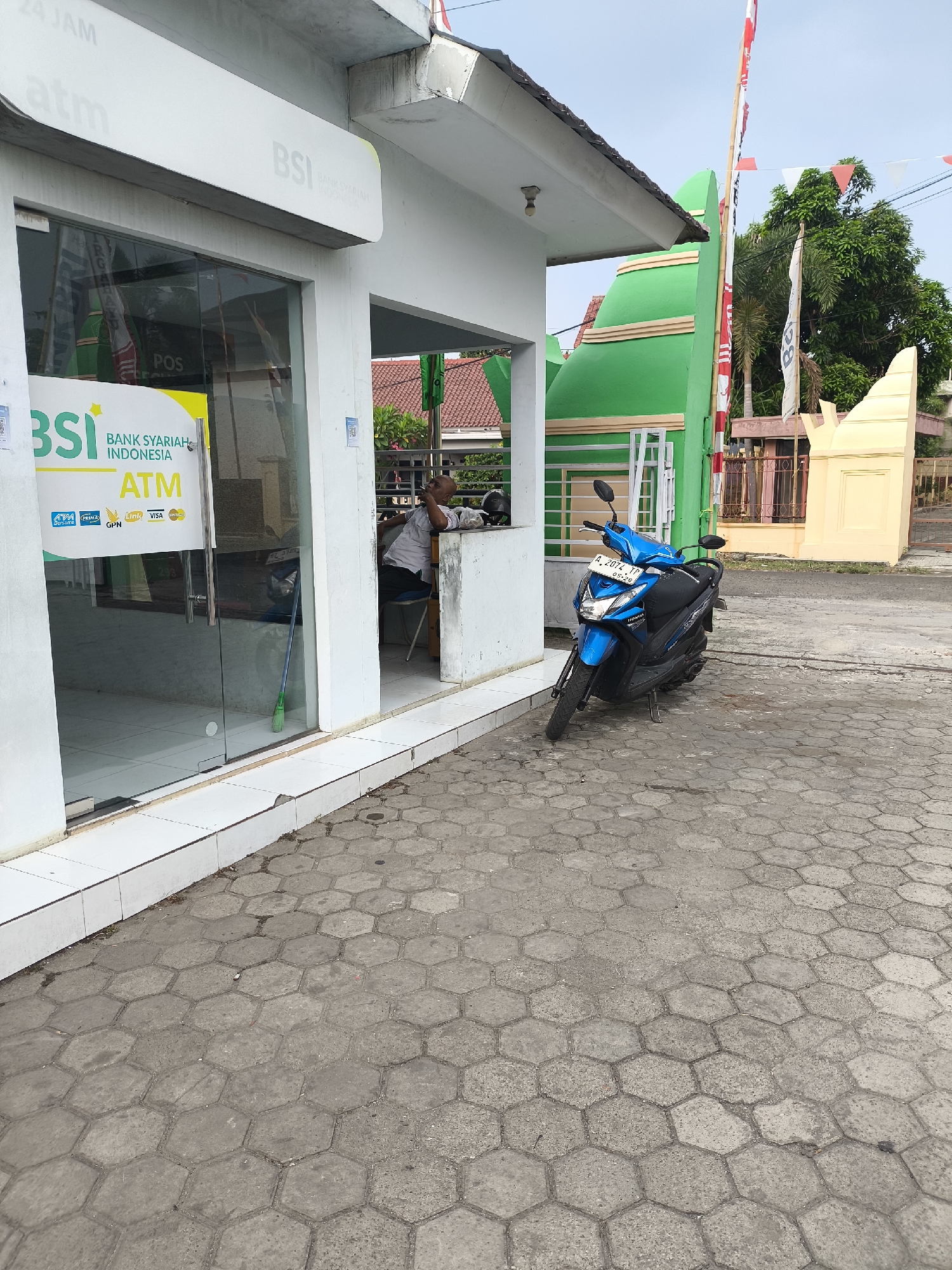 A blue Honda scooter is parked on a hexagonal-paved sidewalk outside a Bank Syariah Indonesia (BSI) ATM booth. The booth has a white facade and a glass door with a BSI ATM sign. Inside the booth, a man in a light-colored shirt and dark pants sits with his head resting on his arm, appearing to be asleep or taking a break. The background features a bright green structure, a flag pole with Indonesian flags, and trees. The scene is set in Cilegon, Indonesia, as indicated by the BSI branding, which is a prominent Islamic bank in Indonesia. The sky is overcast, suggesting it might be a cloudy day.
