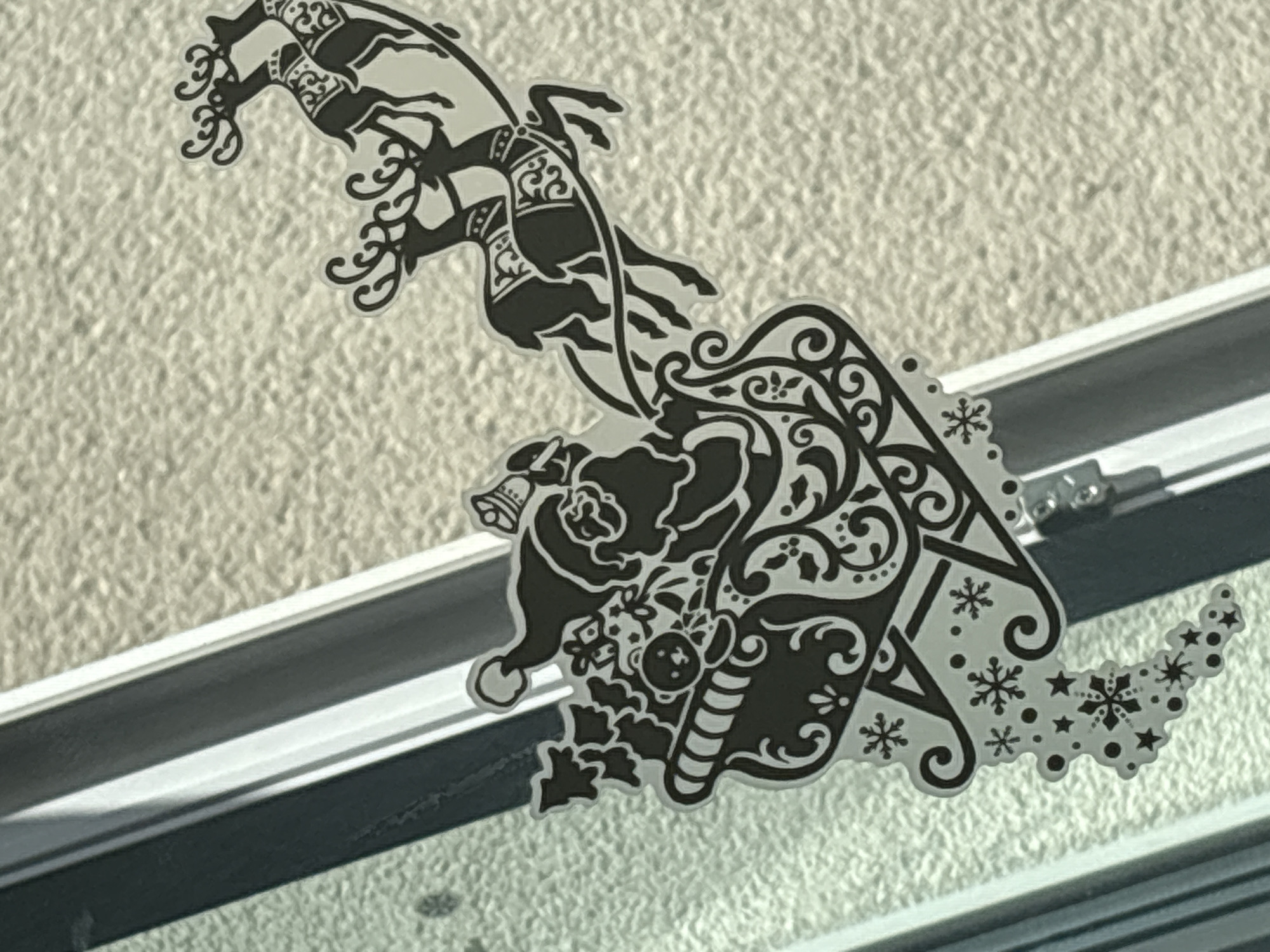 This media file features a black and white decal on a window or glass surface. The decal depicts a whimsical Christmas scene. At the top, two reindeer are shown in mid-gallop, adorned with ornate harnesses and decorative swirls. Below the reindeer, Santa Claus is depicted, wearing his signature hat and coat. He appears to be holding a candy cane and is surrounded by festive elements such as holly, bells, and snowflakes. The overall design is intricate and stylized, with a lot of filigree and decorative patterns integrated into the imagery. The background of the decal is the textured surface of the wall behind the glass. The light suggests it could be daytime, possibly with sunlight casting shadows on the window frame. The window frame itself is white and dark gray, suggesting an indoor setting looking outwards, or an outdoor setting with the decal applied to the exterior of the window. There is no visible text in the image.