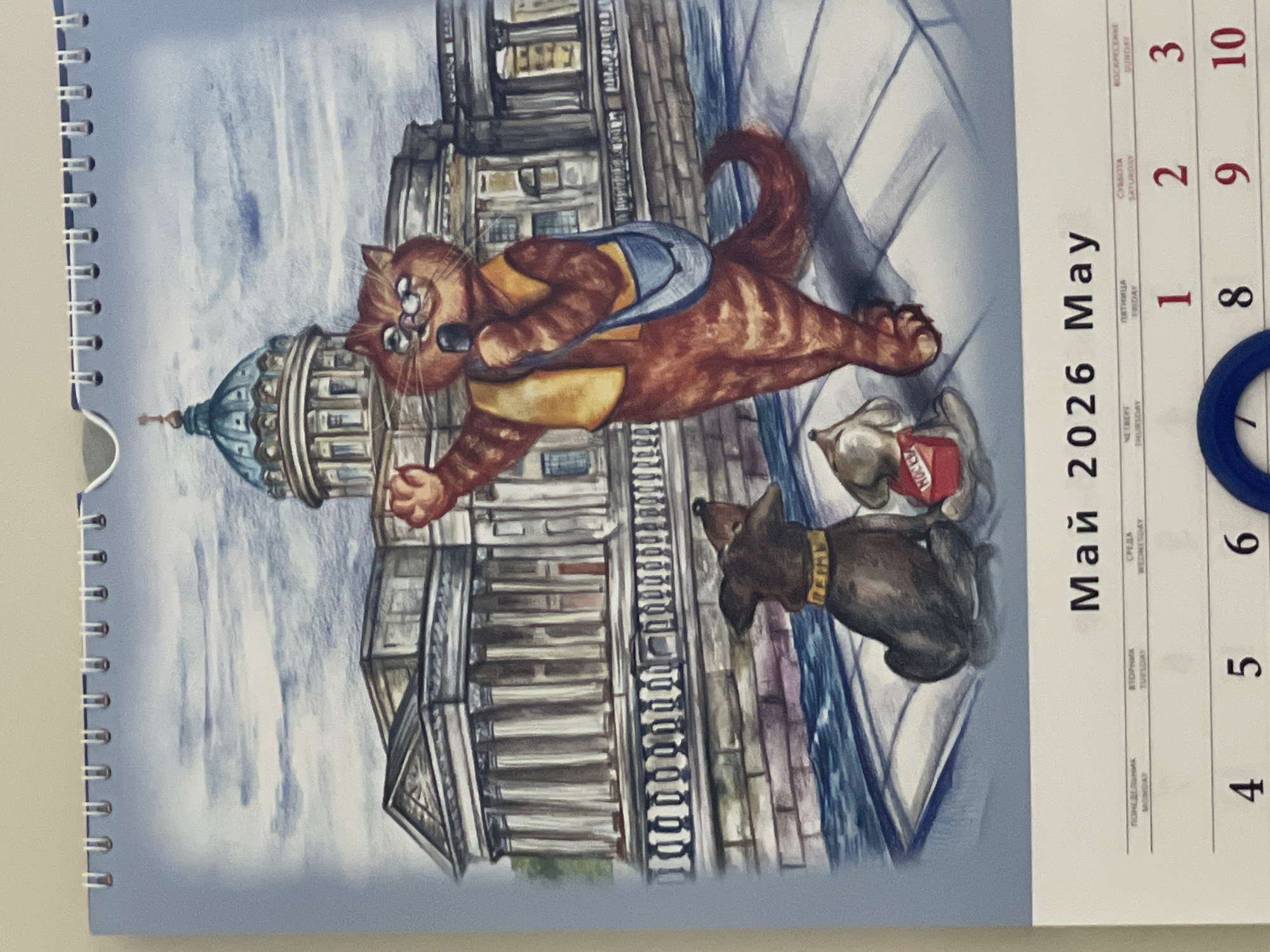 The image is of a May 2026 calendar page with a whimsical illustration. The illustration depicts a red tabby cat wearing a blue and gold backpack, striding purposefully past the Saint Michael's Cathedral in Izhevsk, Russia. A small dark brown dog with a gold collar watches the cat, and a can labeled "MOCKBA" (Moscow) sits on the sidewalk near the dog. The calendar section shows dates from May 1st to 10th, with the 7th highlighted with a blue ring.