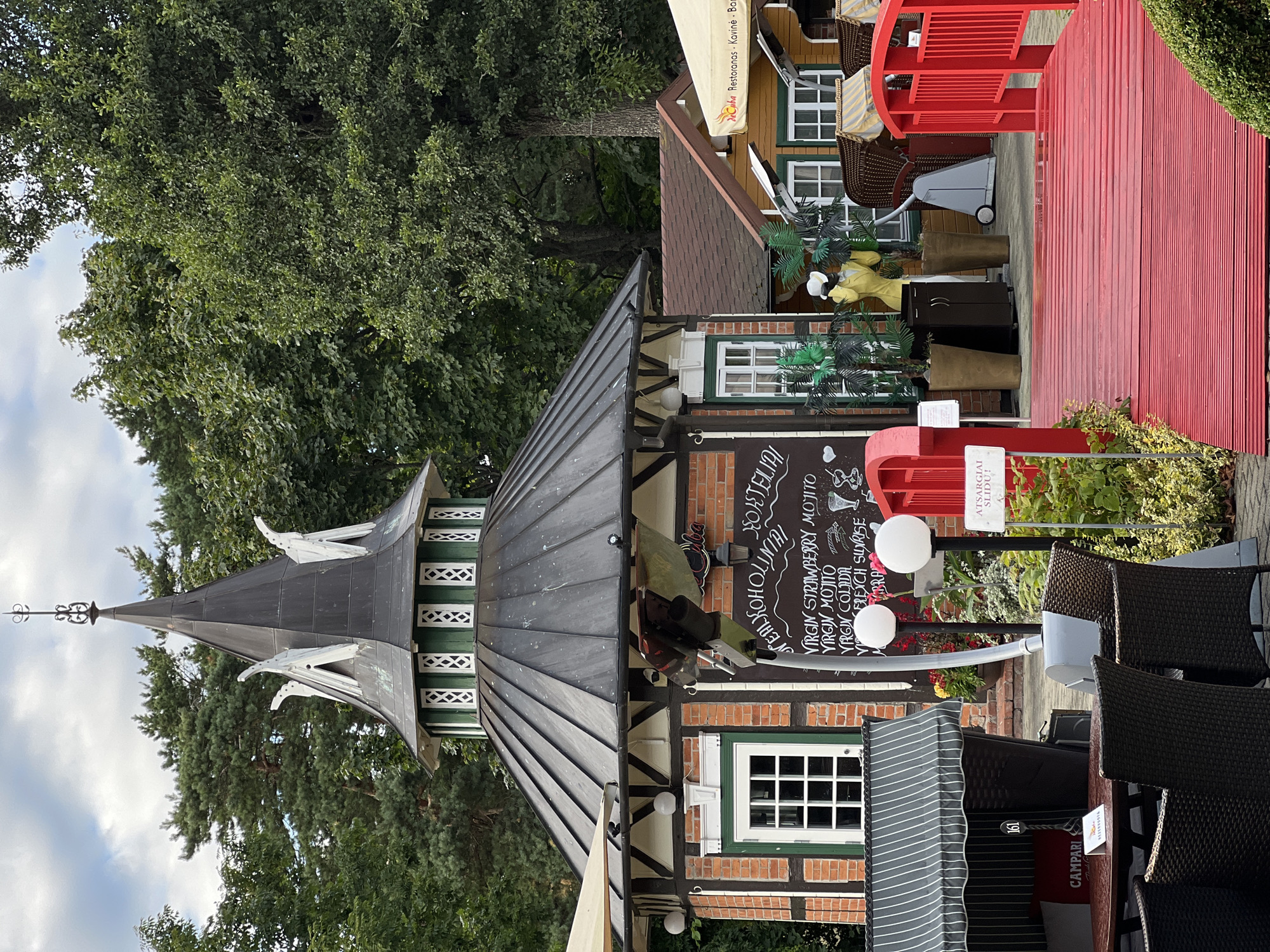 The image depicts an outdoor seating area at a restaurant in Palanga, Lithuania. In the foreground, a bright red bridge with a railing leads to the seating area, which features tables and chairs made of woven material. Some of the tables have "reserved" signs. In the background, there is a building with a distinctive, multi-tiered dark roof and a pointed spire. This building appears to be part of the restaurant, with a brick facade and green and white trim. A chalkboard menu is visible, listing various drinks like "Virgin Strawberry Mojito" and "Virgin Colada." In the middle ground, there are decorative potted plants and two mannequins dressed in yellow, one with a hat. The environment is lush with green trees surrounding the establishment, and the sky is partly cloudy. The scene suggests a pleasant, possibly summer, day at a resort-style dining establishment.