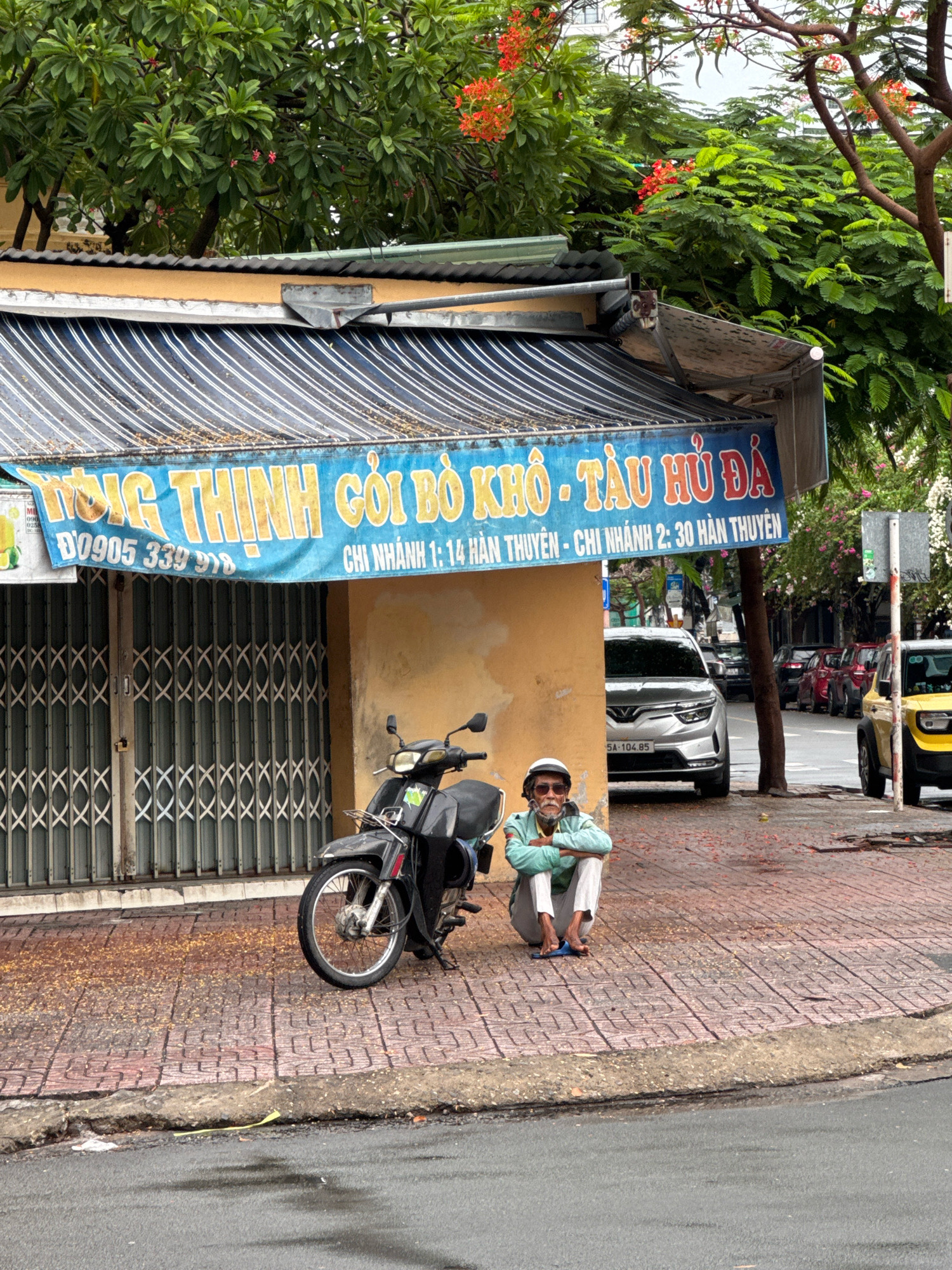 In Nha Trang, Vietnam, on a cloudy day, an elderly man with sunglasses sits on a brick sidewalk near a storefront. He wears a light blue jacket, white pants, and blue flip-flops. A black motorcycle is parked to his left. Above him, a blue awning with yellow lettering stretches across the storefront, advertising "DONG THINH GO BO KHO - TAU HU DA" and providing contact information. The storefront is covered by a metal accordion gate. The street in front of the storefront is lined with parked cars, including a silver sedan and a yellow SUV. Trees with green leaves and some red flowers are visible behind the awning and along the street. The sidewalk is littered with fallen leaves and debris.