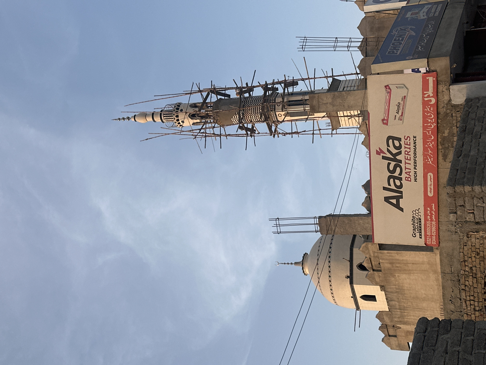 The image captures a partially constructed minaret and dome of a mosque in Quetta City Tehsil, Pakistan. The minaret is surrounded by scaffolding, indicating ongoing construction. A large billboard advertising "Alaska Batteries" is prominently displayed in the foreground, along with another billboard for "Phoenix Batteries". The sky above is clear with some wispy clouds. The architectural style suggests a traditional Islamic design. The scene appears to be in an urban or semi-urban setting.