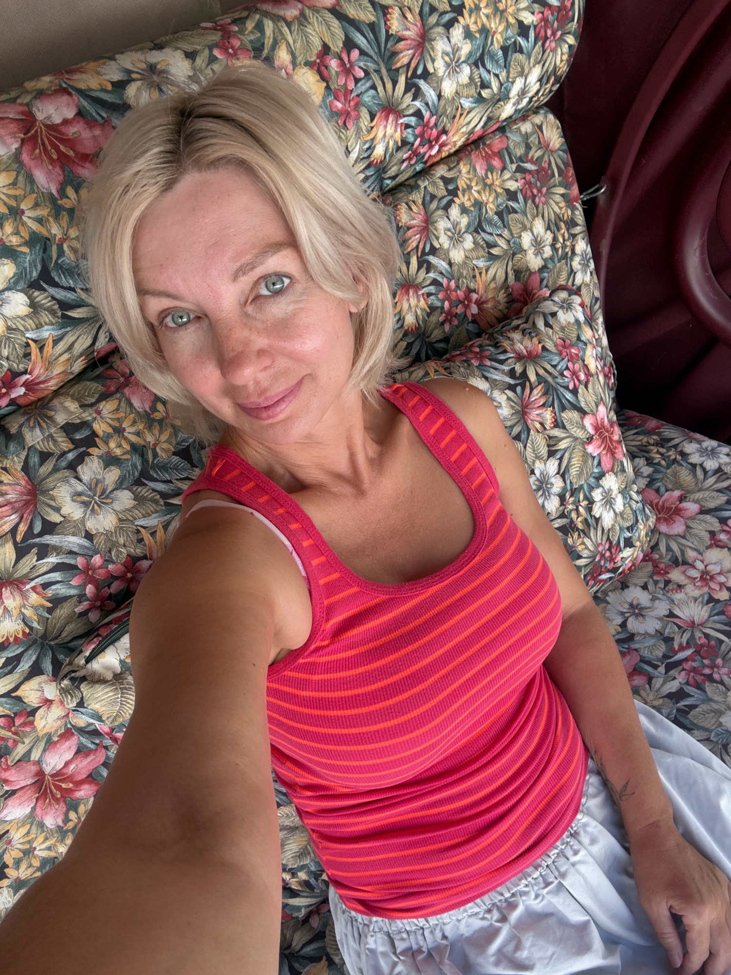A woman with blonde hair, blue eyes, and fair skin, wearing a pink and orange striped tank top and light-colored pants, is lying down on a floral-patterned cushion. She is looking at the camera and smiling slightly. The setting appears to be outdoors, possibly a patio or garden area, with a dark red curved object, likely part of a piece of furniture such as a swing or chair, visible in the background. The floral pattern on the cushions features a variety of flowers in shades of pink, red, yellow, and green against a dark background. The image is taken from a high angle, looking down at the woman. No specific landmarks in Abrau-Dyurso, Russia are identifiable from the image alone.