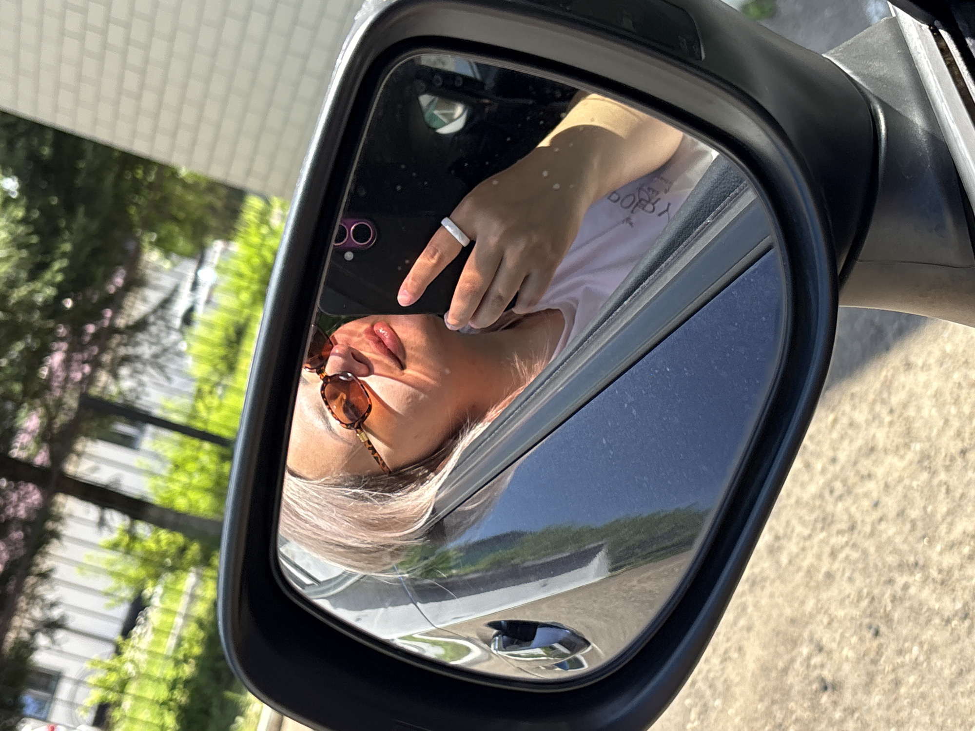 A woman with blonde hair and sunglasses is taking a selfie in a car's side-view mirror. She is wearing a white t-shirt and a ring on her left hand. The background of the mirror reflection shows trees and a building. The car is parked on a street in Birobidzhan, Russia, evidenced by the text on the woman's t-shirt. The overall scene suggests a sunny day.