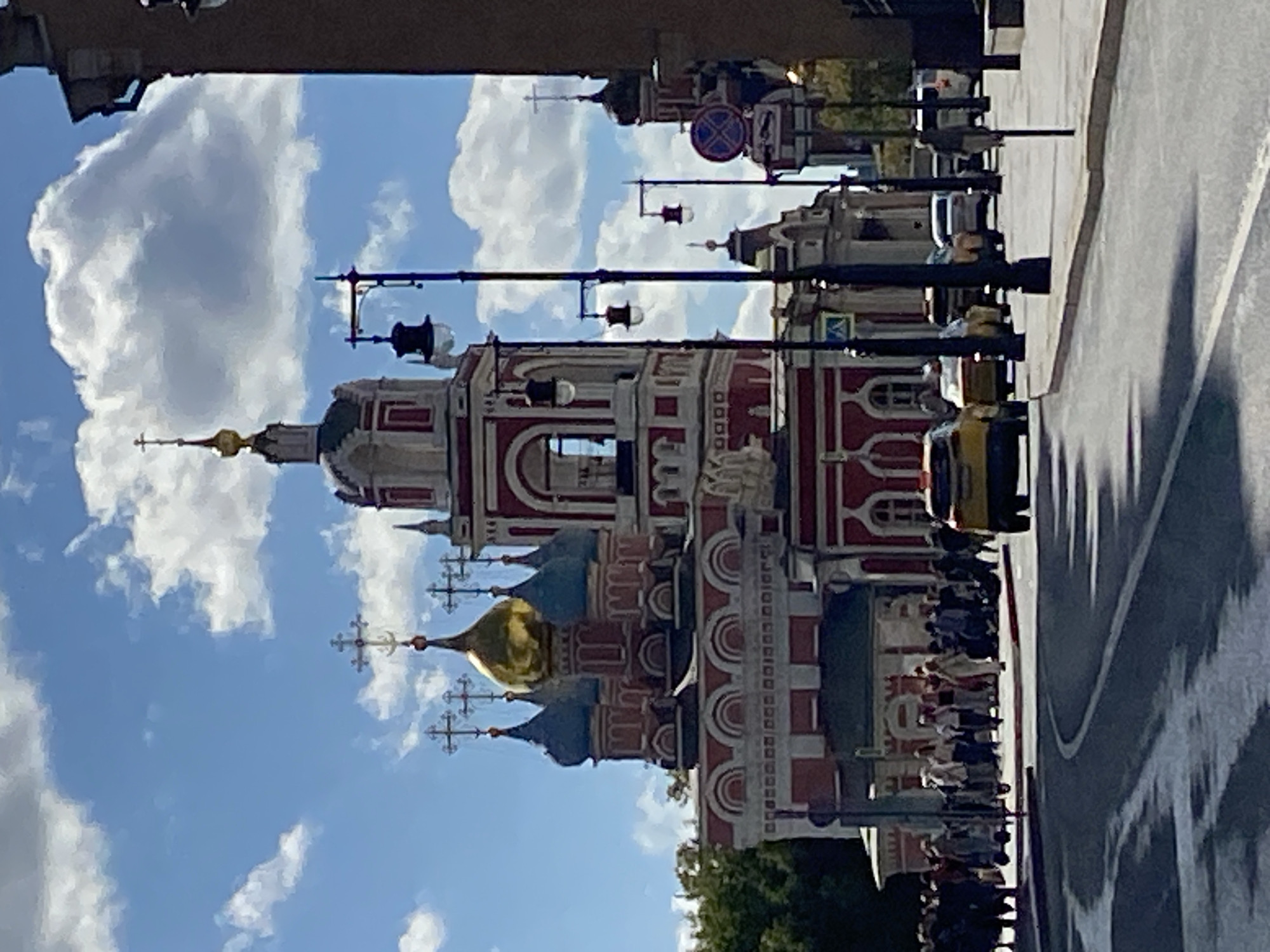 The image depicts a street scene in Moscow, Russia, with a prominent red and white church featuring multiple domes and crosses in the background. Several cars are visible on the road, including a yellow taxi. A group of people are walking on the sidewalk in front of the church. The sky is blue with scattered white clouds. Lampposts line the sidewalk. The scene appears to be a sunny day.