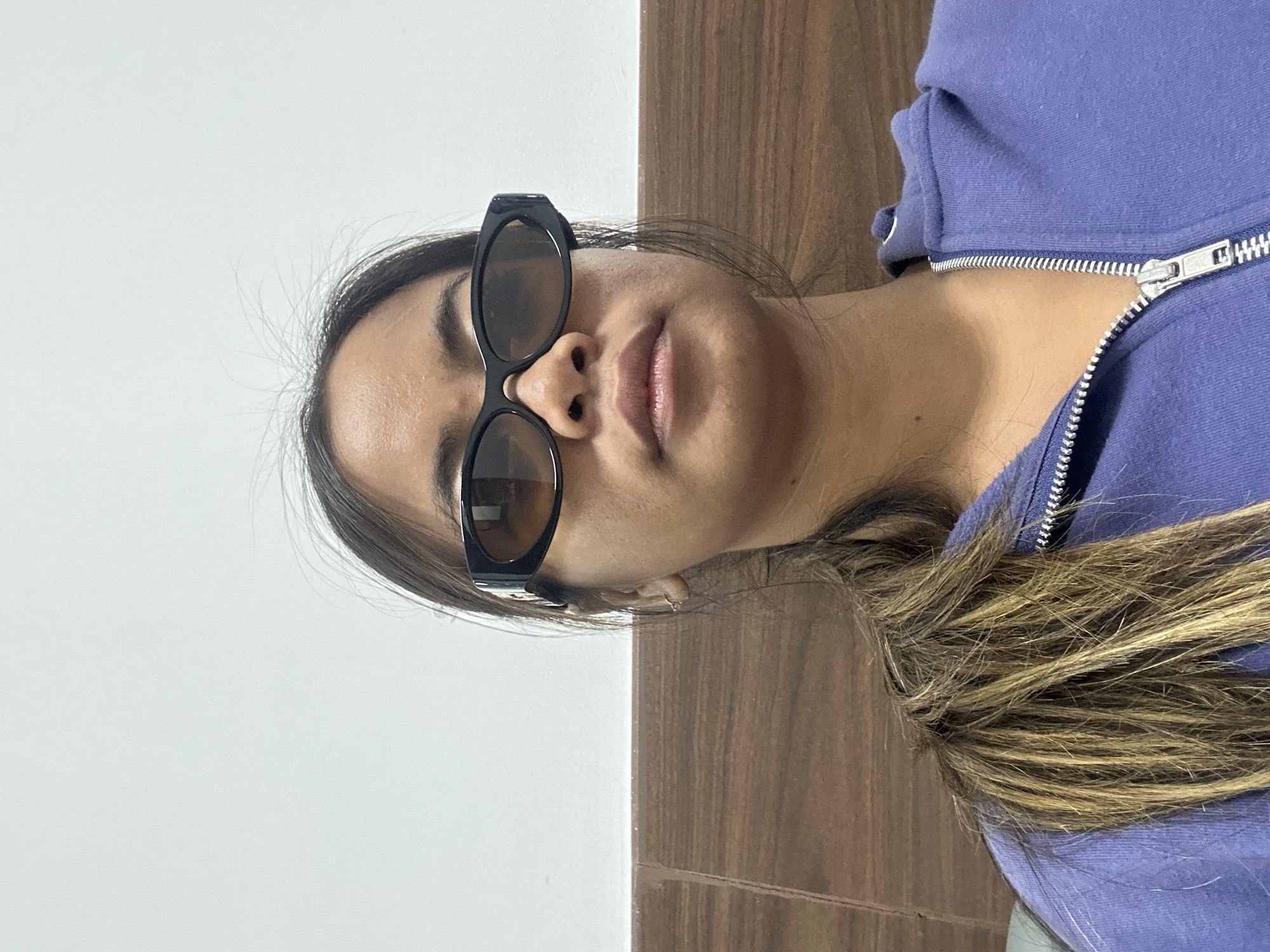 A young woman with dark hair pulled into a ponytail and wearing black sunglasses and a purple hooded jacket is shown in a close-up, frontal shot. She is looking directly at the camera. The background consists of a white wall above a wooden panel. The scene is likely indoors, possibly in a hotel room or similar accommodation in Kuta, Indonesia, given the context of the request.