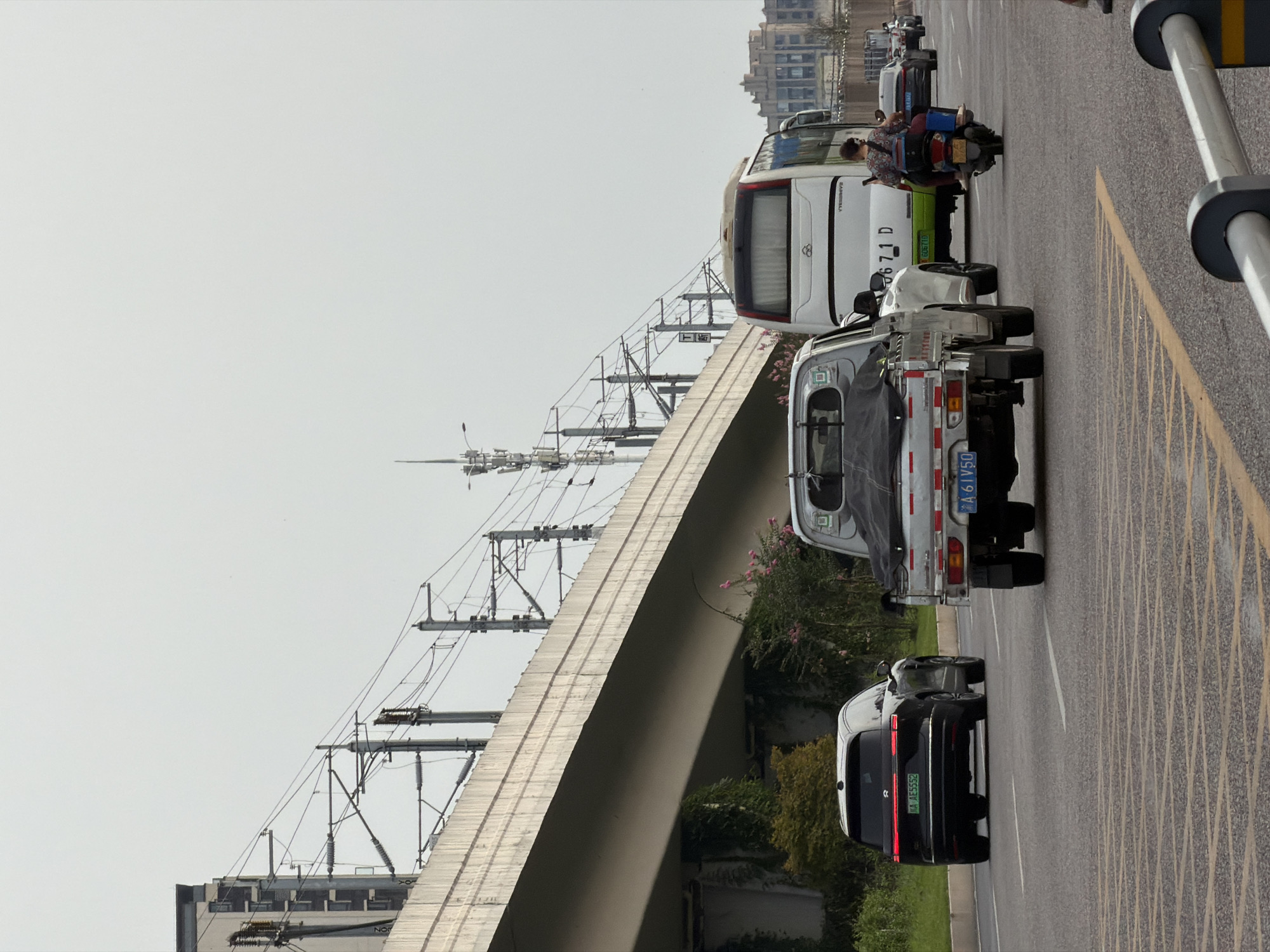 A medium shot captures a traffic scene on a cloudy day, likely in the city of Jiangjin, China. In the foreground, a white pickup truck with a tarp-covered bed is moving forward. To its left, a black SUV with a red taillight bar is also in motion. Behind the pickup truck, a white bus with a green license plate is visible. A person on a scooter, wearing a patterned shirt and dark pants, is riding in front of the bus. In the distance, a multi-story building with a patterned facade stands out against the overcast sky. The scene is framed by overhead power lines and a concrete overpass, suggesting an urban or suburban setting with elevated infrastructure. The road is asphalt, marked with yellow and white lines.