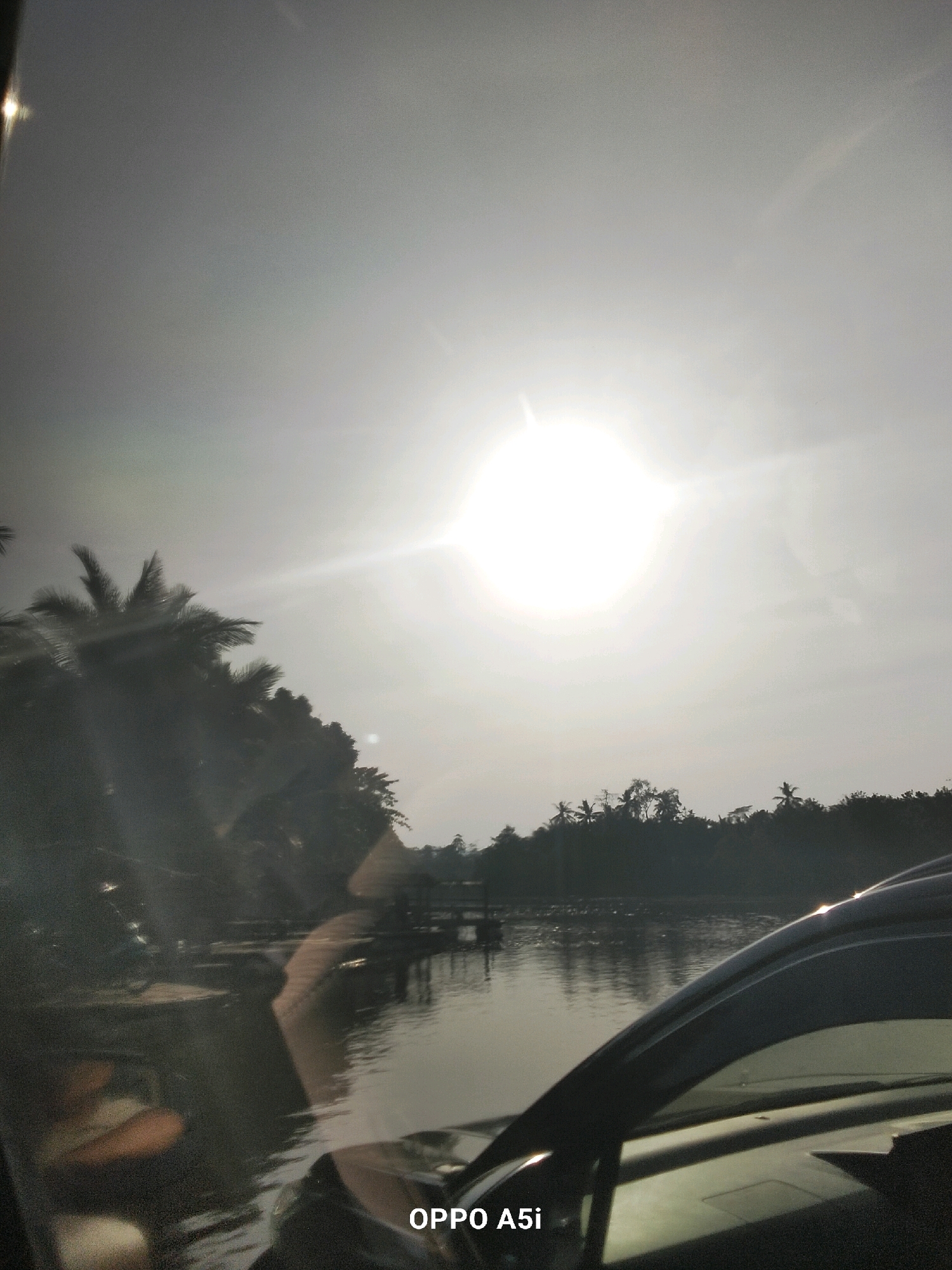 The image captures a bright, hazy sun shining over a body of water, likely a river or lake, bordered by lush vegetation. The sun's glare is intense, creating lens flares and washing out much of the detail in the sky. On the left side of the frame, silhouetted palm trees and other dense foliage are visible. In the foreground, the reflection of trees and the sky can be seen on the water's surface. The lower right corner of the image shows a portion of a dark-colored vehicle, possibly a car, with its side mirror and window reflecting the scene. The bottom edge of the image contains the text "OPPO A5i", indicating the device used to capture the photograph. The scene appears to be a natural landscape, possibly a rural or semi-rural area, given the vegetation and the body of water. The lighting suggests it is daytime, with the sun high in the sky. The general atmosphere is one of a bright, natural setting. The location is identified as Tabanggele, Indonesia.