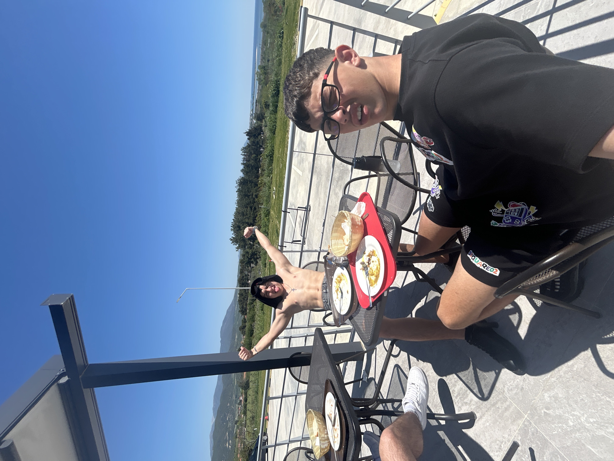 Two young men are posing on a rooftop terrace in Νέα Κερδύλια, Greece. One man is shirtless, wearing a black hoodie, and has his arms raised in a celebratory gesture. The other man, closer to the camera, is wearing a black t-shirt and shorts, both with graphic designs, and glasses. They are seated at a metal table with a red tray holding plates with food remnants and a bread basket. In the background is a landscape with trees, mountains, and a coastline. The sky is clear blue.
