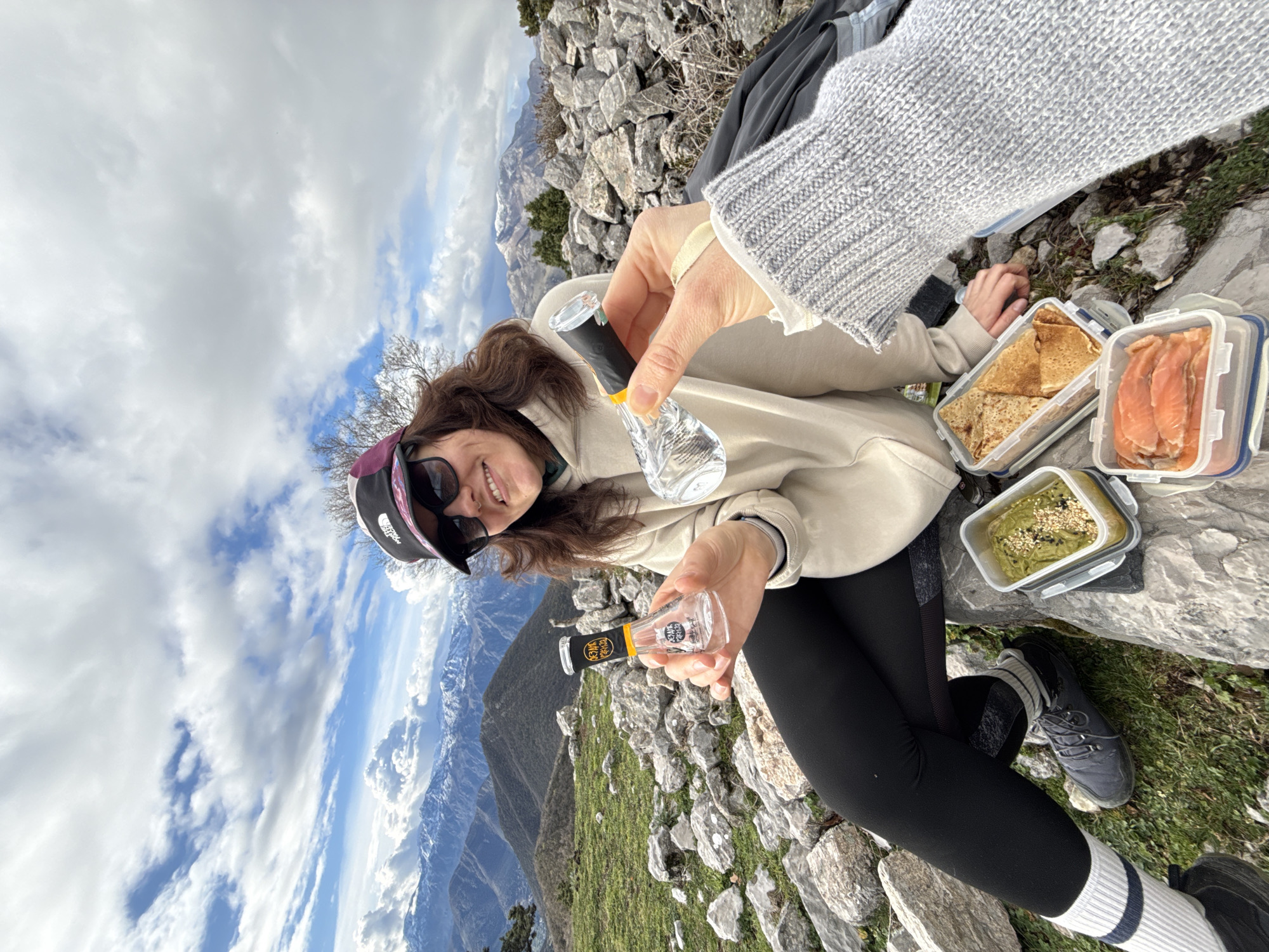 The scene depicts two individuals at a high-altitude mountainous vantage point near Baošići, Montenegro. The primary subject, a female, is seated on a rock, smiling at the camera. She is wearing a dark baseball cap, sunglasses, a light-colored hooded sweatshirt, black leggings, white socks with dark stripes, and dark hiking shoes. She holds a small, clear, flask-shaped bottle labeled "ОГЊЕНА РАКИЈА" in her left hand. The second individual's right arm, wearing a grey knitted sweater, extends into the frame, holding a similar clear, uncapped bottle. Adjacent to the seated individual are three clear plastic food containers with blue lids: one contains a green, textured spread topped with seeds, another holds folded crêpes, and a third contains slices of smoked salmon. The immediate environment consists of grassy patches interspersed with numerous light-colored rocks. In the background, snow-capped mountain ranges are visible under a partly cloudy sky with patches of blue.