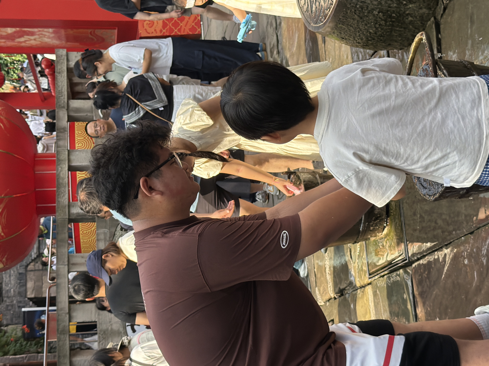The image shows a group of people in an outdoor setting that appears to be a public area, possibly a market or a tourist spot in Yuzhong District, China, as indicated by the architectural style and red decorative elements suggestive of Chinese culture. In the foreground, a man wearing a brown t-shirt is leaning down, interacting with a young child who is facing away from the camera, wearing a white t-shirt. The man's hands are near a dark bucket, and the child's hands are also near a bucket. Behind them, other people are visible, including a woman in a white t-shirt and dark pants, and a child in a light-colored dress holding a blue water gun. The ground is wet and appears to be paved with stone or concrete. In the background, there is a red structure with decorative patterns, possibly a building or a temporary stage. People are seen milling about, suggesting a lively atmosphere. The overall lighting suggests it is daytime.
