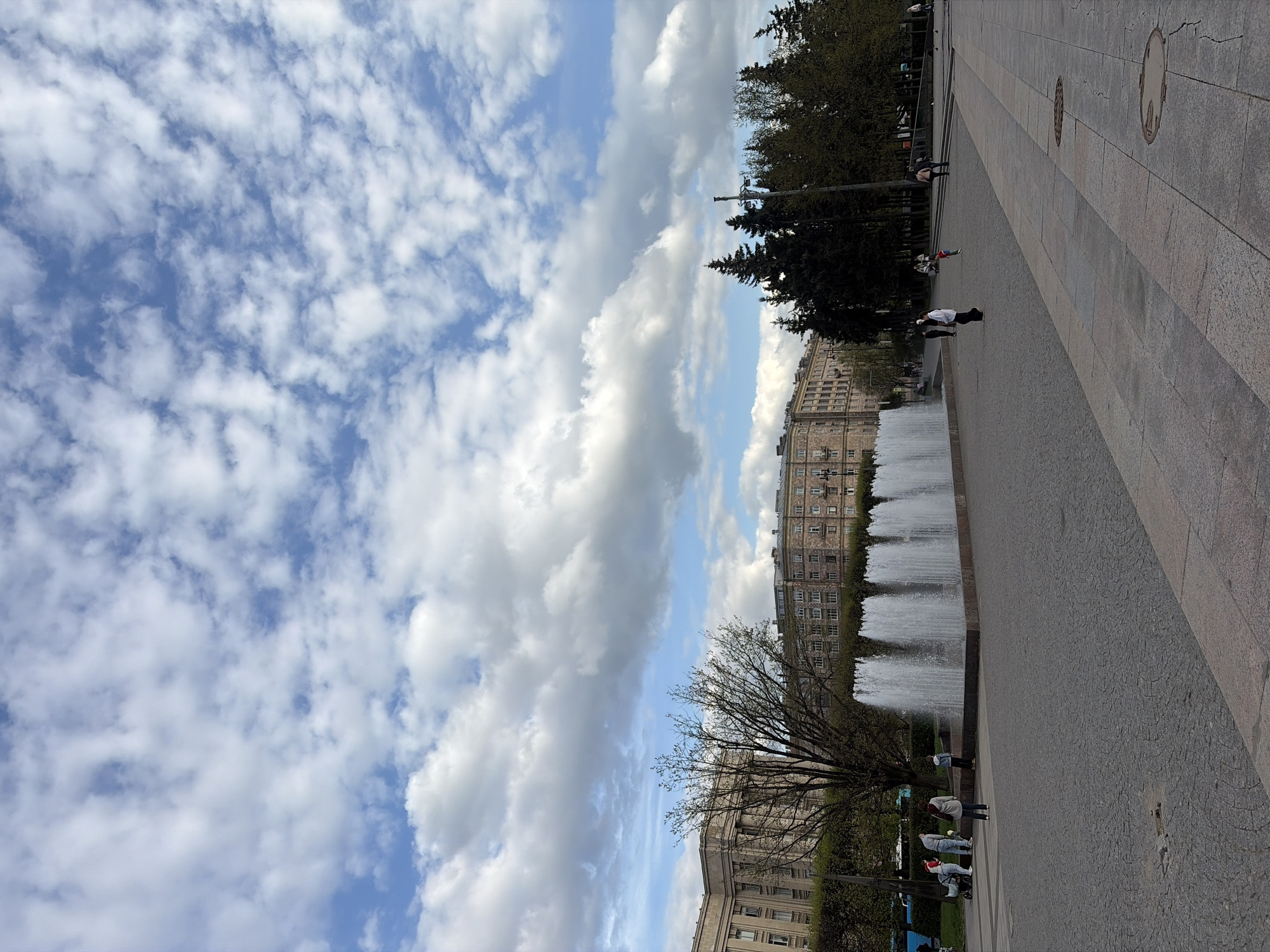 The image captures a view of a city landscape in Saint Petersburg, Russia. The sky is filled with puffy white clouds against a blue background. A wide pedestrian walkway takes up much of the foreground and right side of the image.
A row of tall, dark green trees lines the edge of the walkway along the median, with buildings visible behind them. A fountain with multiple streams of water is present on the right side of the image, along the building's edge. People can be seen walking or standing along the walkway near the fountain and trees.
The buildings are multi-story and appear to be residential or office structures. The overall scene suggests a peaceful, urban environment with a combination of natural and man-made elements.